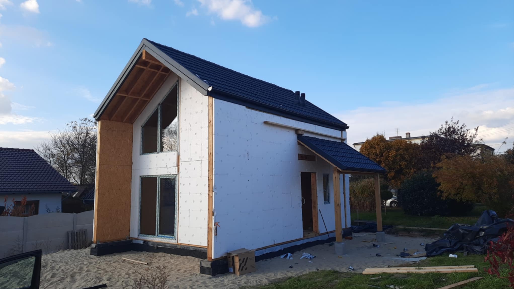 Niedokończony dom modułowy z drewnianą konstrukcją szkieletową i ociepleniem styropianowym, widoczne elementy dachu pokrytego ciemną dachówką, teren budowy z piaskiem i narzędziami.