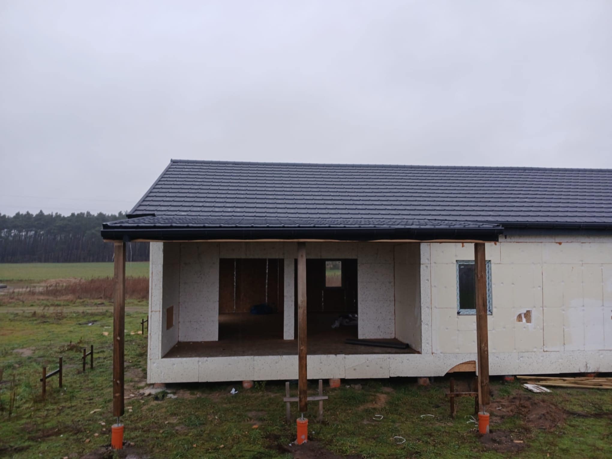 Surowy stan domu modułowego z szarą dachówką, drewnianymi słupami i niezabudowanymi otworami okiennymi na tle pochmurnego nieba i zielonego terenu.