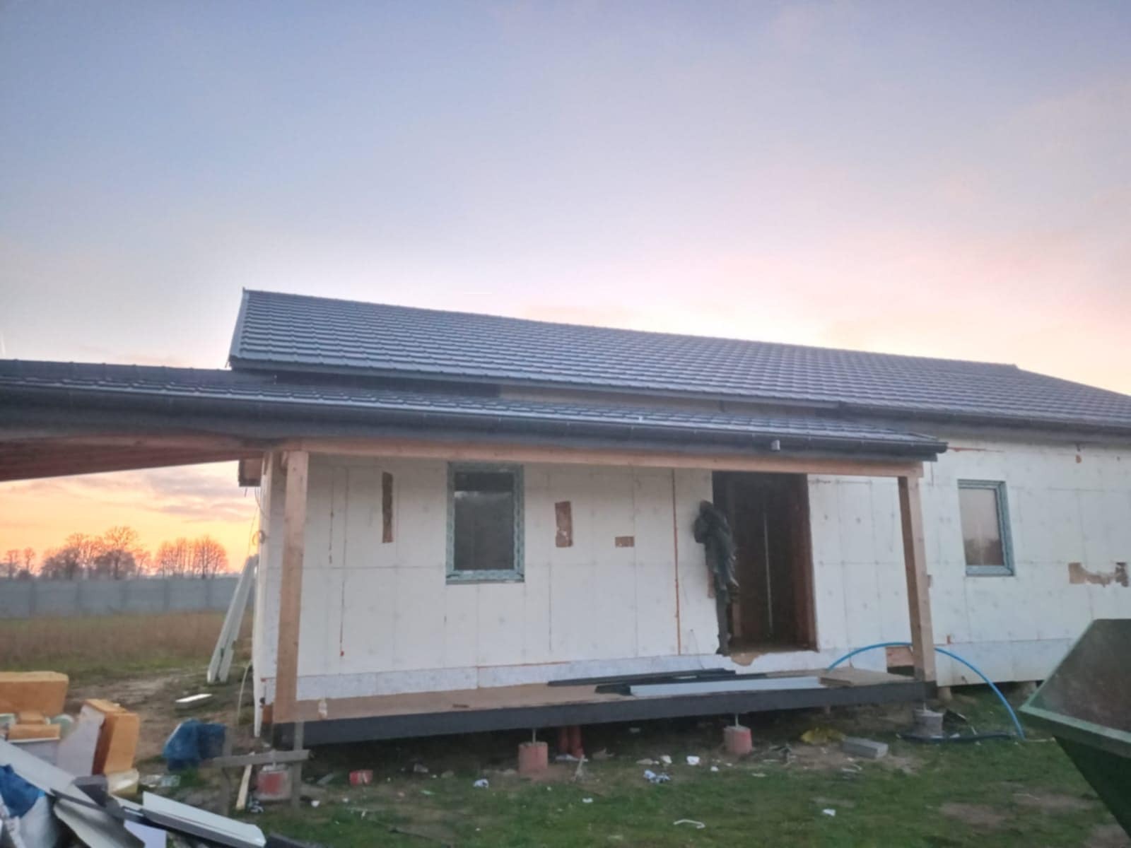Wznoszenie domu modułowego z widoczną izolacją ścian zewnętrznych i fragmentem dachu pokrytego szarą dachówką, na tle zachodzącego słońca.