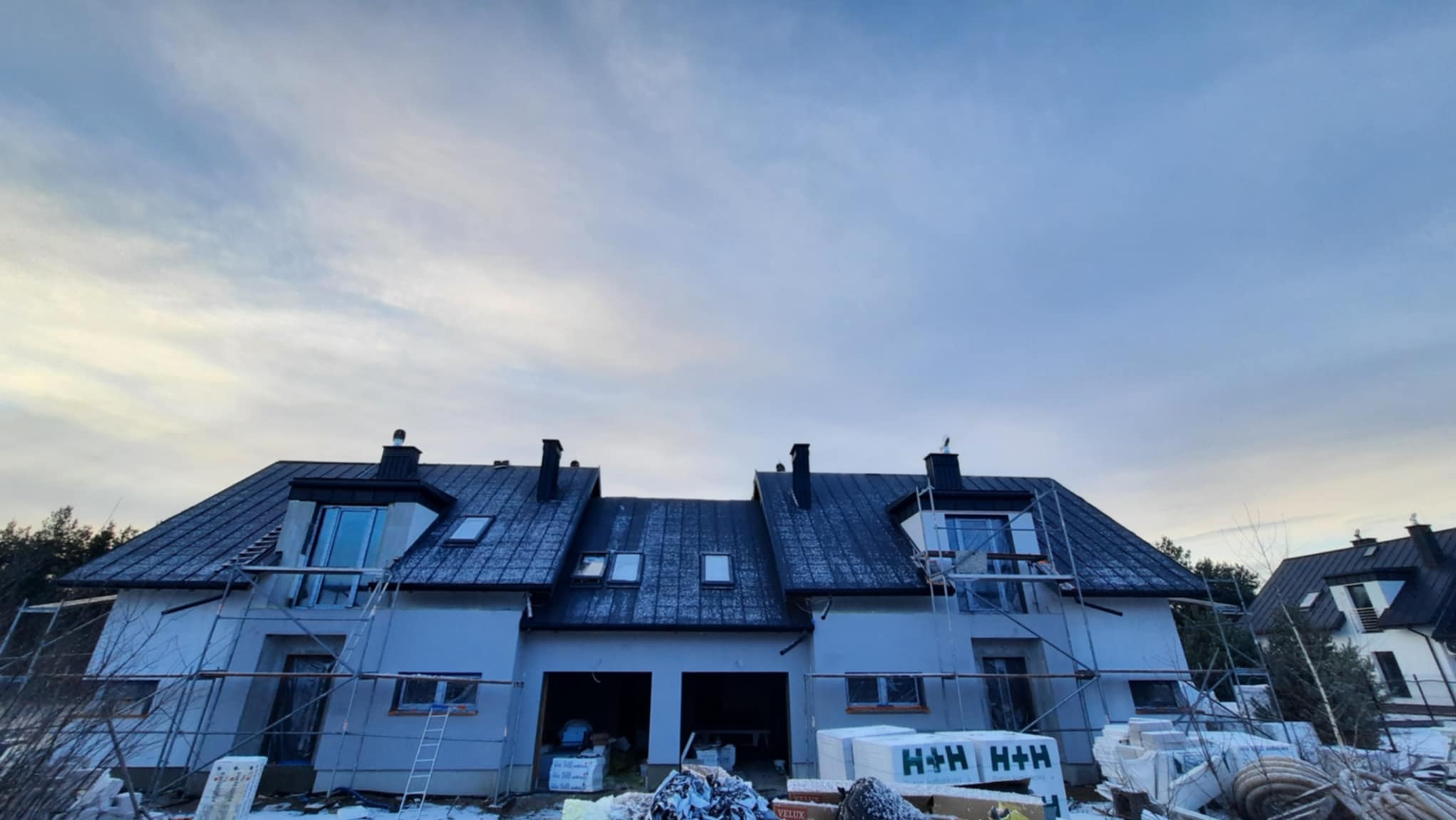 Domy w zabudowie szeregowej w trakcie budowy, z czarnym dachem i widocznymi rusztowaniami, materiały budowlane na pierwszym planie, pochmurne niebo w tle.