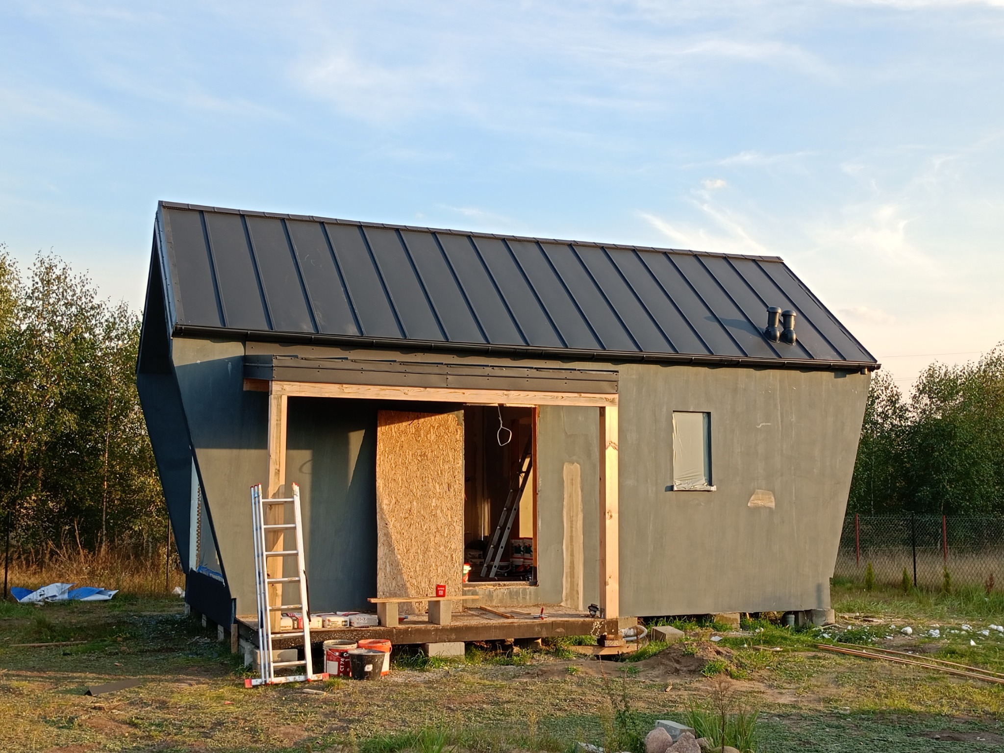 Niedokończony dom modułowy z czarnym dachem z blachy, widoczna drabina, materiały budowlane i surowe drewniane elementy konstrukcyjne.