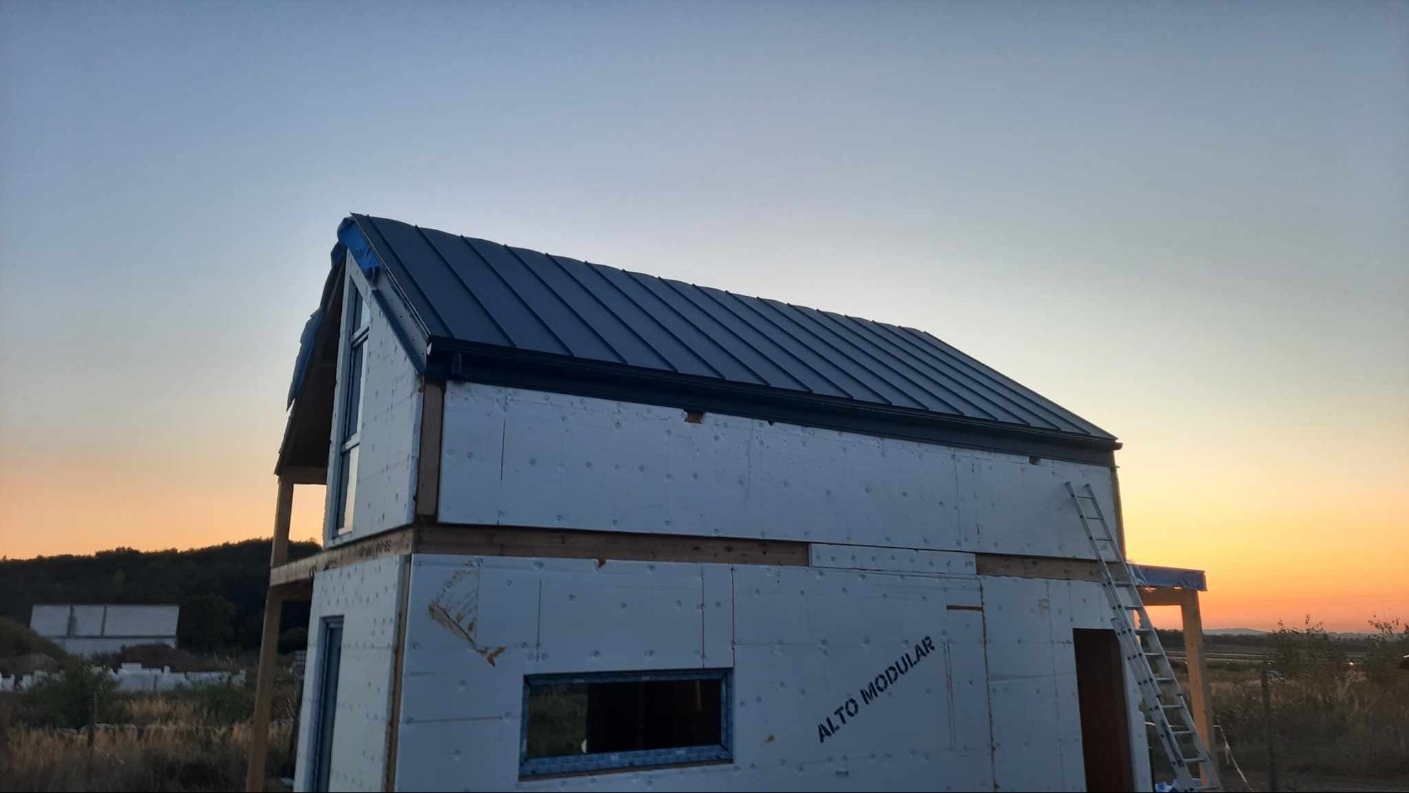 Wznoszony dwukondygnacyjny dom modułowy z ciemnym dachem, widoczna drabina oparta o ścianę, na elewacji widnieje napis ALTO MODULAR, w tle zachód słońca.