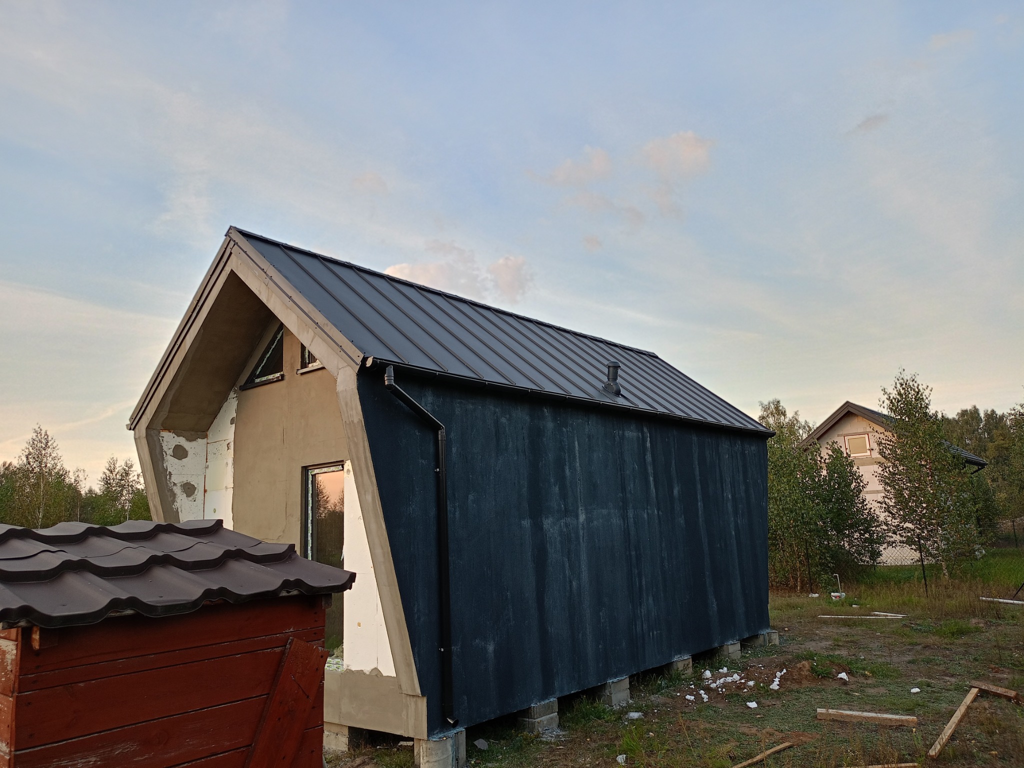 Niedokończony dom modułowy z czarną elewacją i grafitowym dachem, widoczny styropian i surowy beton, w tle drugi dom i drzewa.