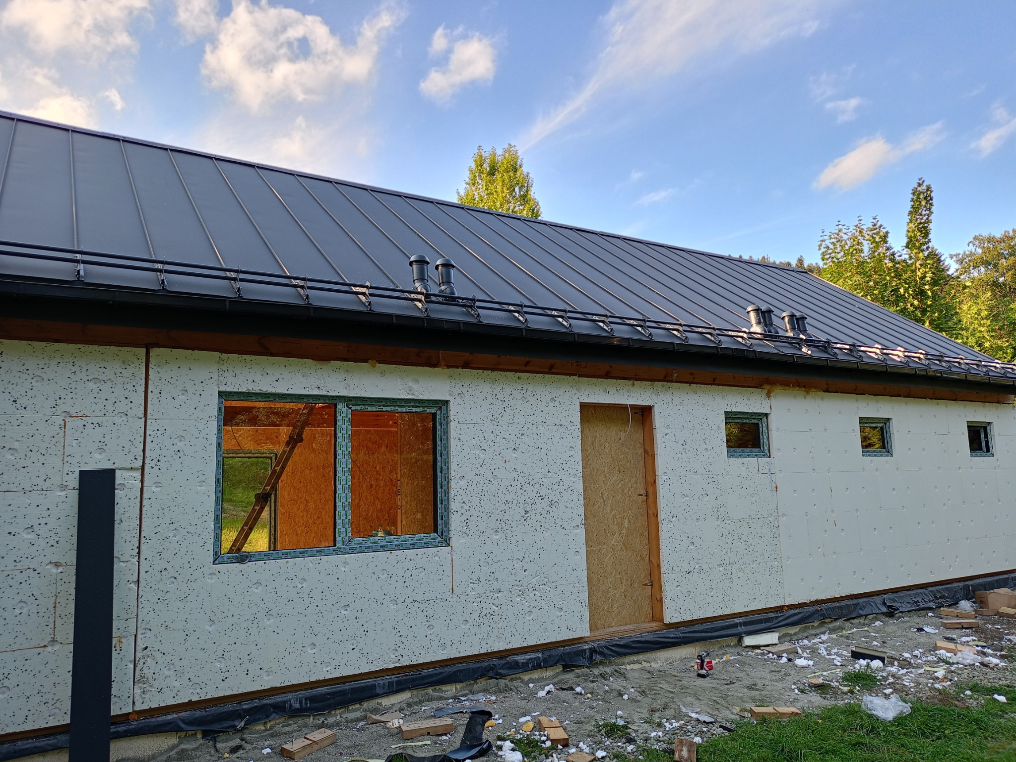 Budynek w trakcie budowy, ocieplony styropianem, z zamontowanymi oknami i drzwiami, pokryty blachą trapezową w kolorze grafitowym, widoczne rynny i kominki wentylacyjne.