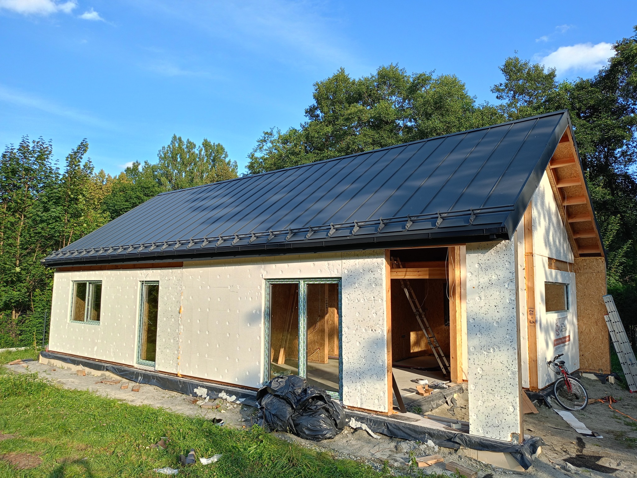 Niedokończony dom modułowy z ciemnoszarym dachem, widoczne elementy konstrukcyjne, styropianowe ocieplenie ścian, okna z zielonymi ramami oraz rower i drabiny w tle