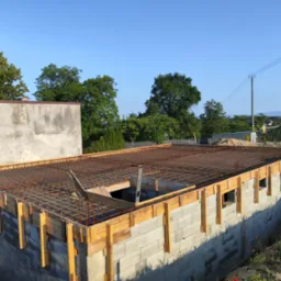 Wznoszenie ścian z bloczków betonowych z deskowaniem i zbrojeniem pod wylewkę stropu, widok z góry na plac budowy w słoneczny dzień.