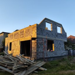 FRB ORAWIEC Wojciech Orawiec - Surowy stan domu z bloczków, wczesnym rankiem, z widocznymi rusztowaniami i stertą desek na pierwszym planie, przy wschodzącym słońcu.
