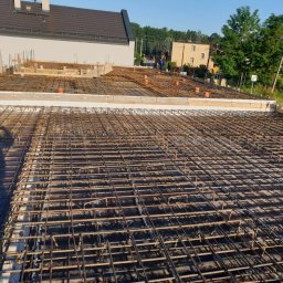 FRB ORAWIEC Wojciech Orawiec - Zbrojenie płyty fundamentowej z widocznymi elementami szalunku i wystającymi prętami, w tle zabudowa jednorodzinna i zieleń.