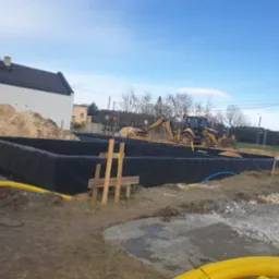 Żółta koparka zasypuje piaskiem wykop pod budynek, zabezpieczony czarną folią. Widoczne wystające rury i drewniane stemple. W tle dom i drzewa.