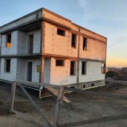 Surowy dwukondygnacyjny budynek z białych bloczków w Bieruniu, widoczna izolacja fundamentów i tymczasowe ogrodzenie na tle zachodzącego słońca.