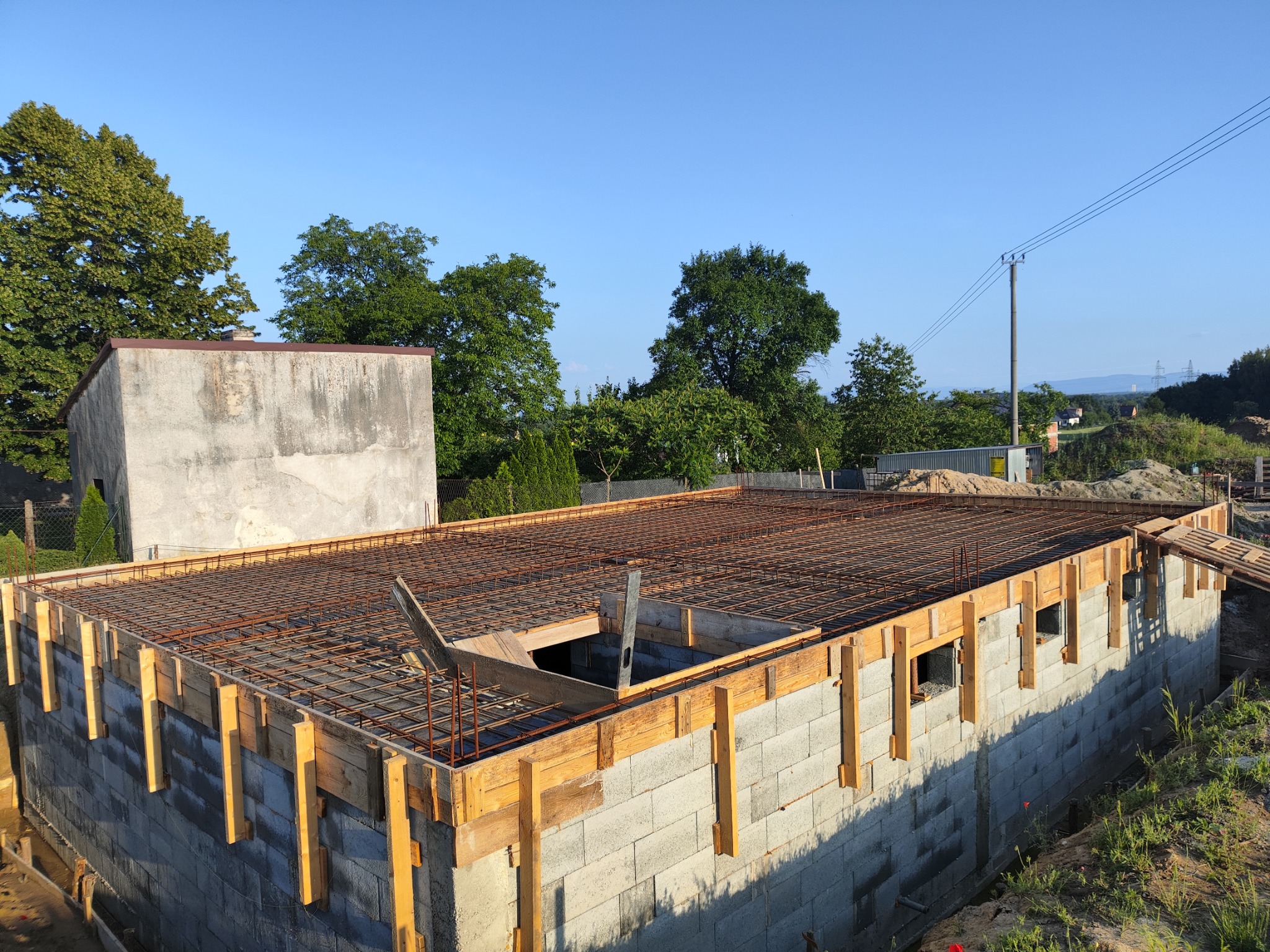 Wznoszenie ścian z bloczków betonowych z deskowaniem i zbrojeniem pod wylewkę stropu, widok z góry na plac budowy w słoneczny dzień.