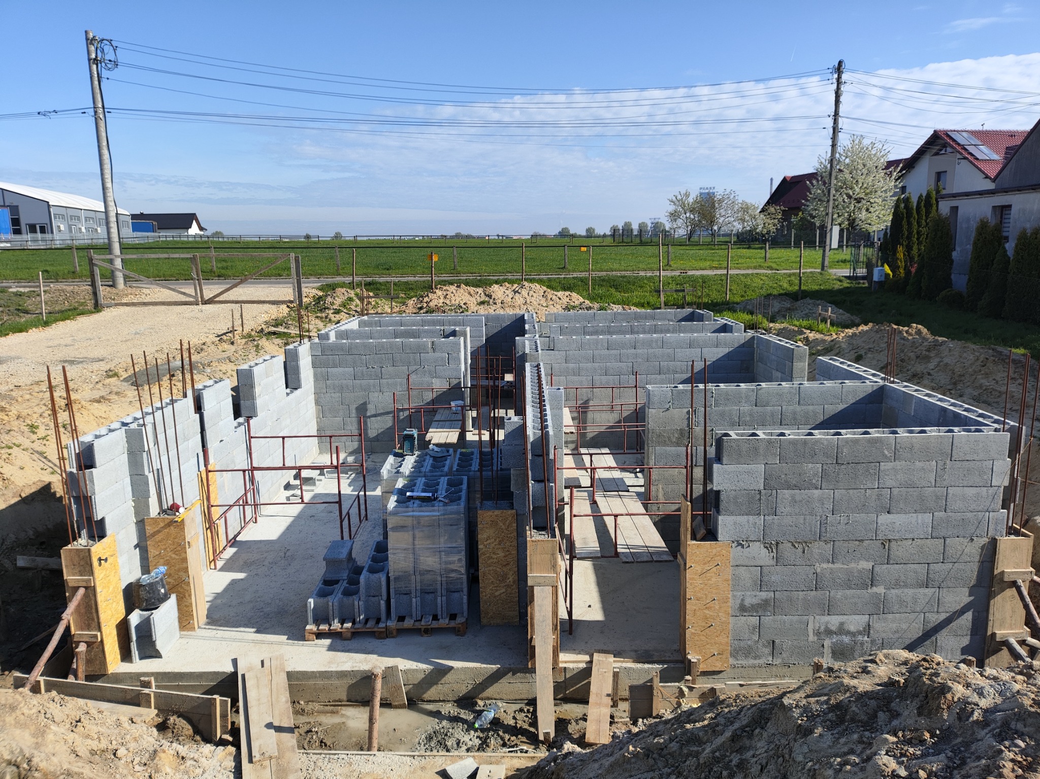 Wznoszenie ścian fundamentowych z bloczków betonowych na budowie domu jednorodzinnego, widoczne zbrojenie i szalunki, w tle krajobraz wiejski pod błękitnym niebem.