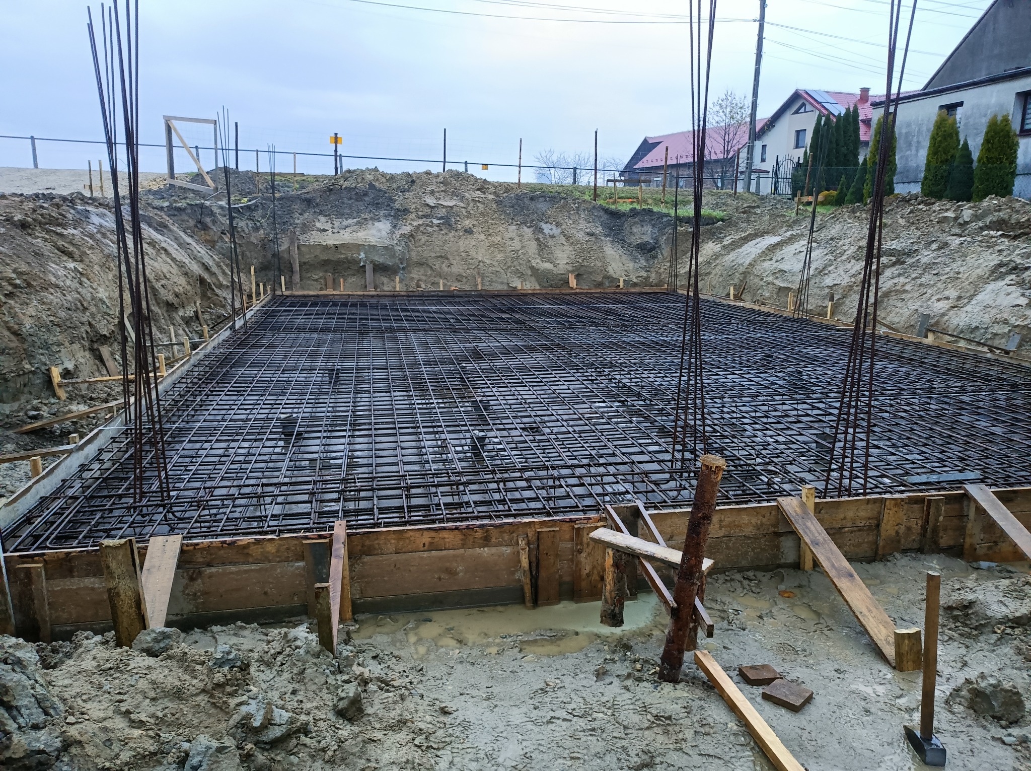 Zbrojenie fundamentu w wykopie, drewniany szalunek, wystające pręty zbrojeniowe, widok na teren budowy i zabudowania w tle.