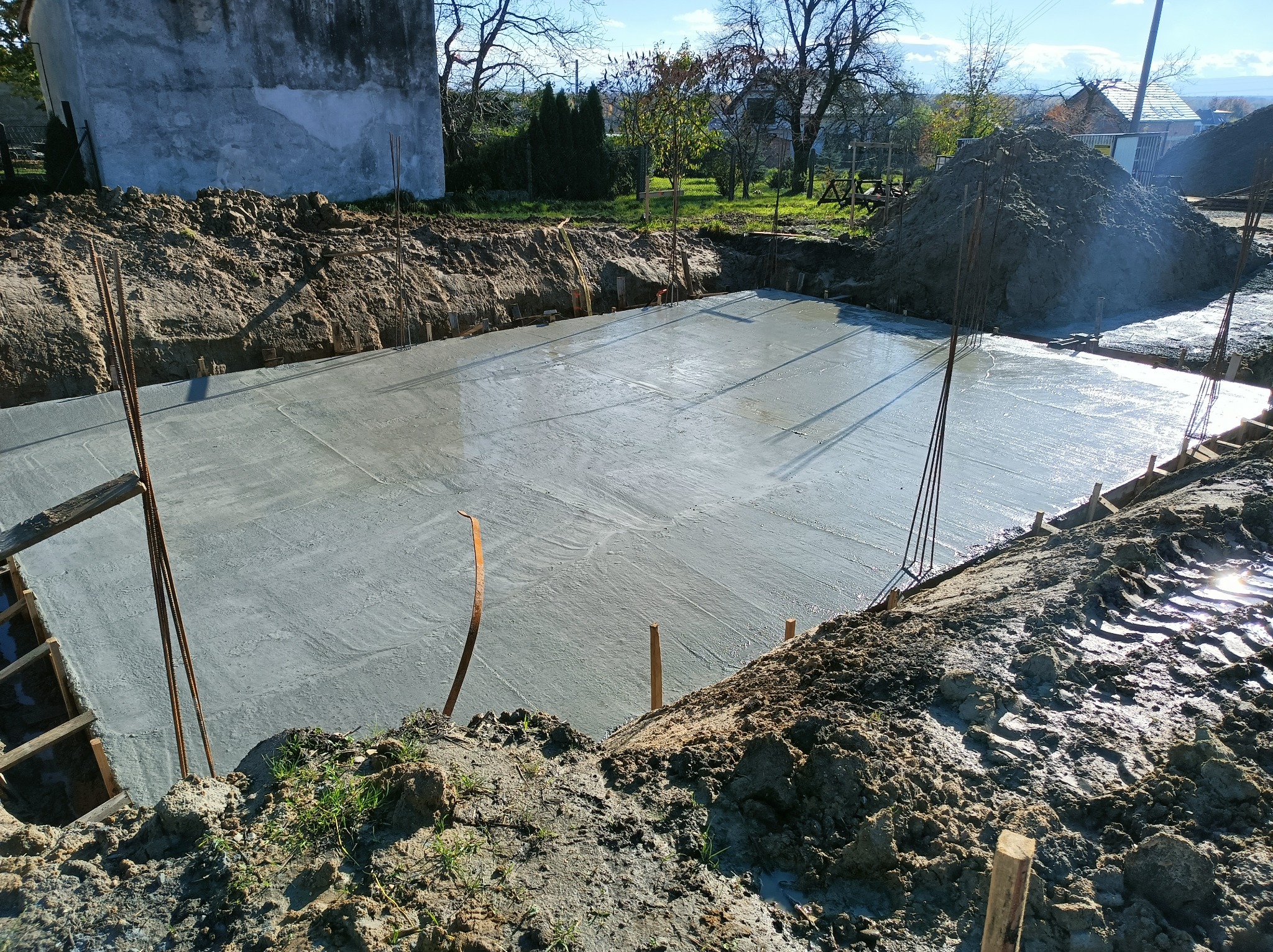 Świeżo wylana, gładka płyta fundamentowa w wykopie, z wystającymi prętami zbrojeniowymi i drewnianym szalunkiem, otoczona stertami ziemi i widokiem na zabudowania w tle.