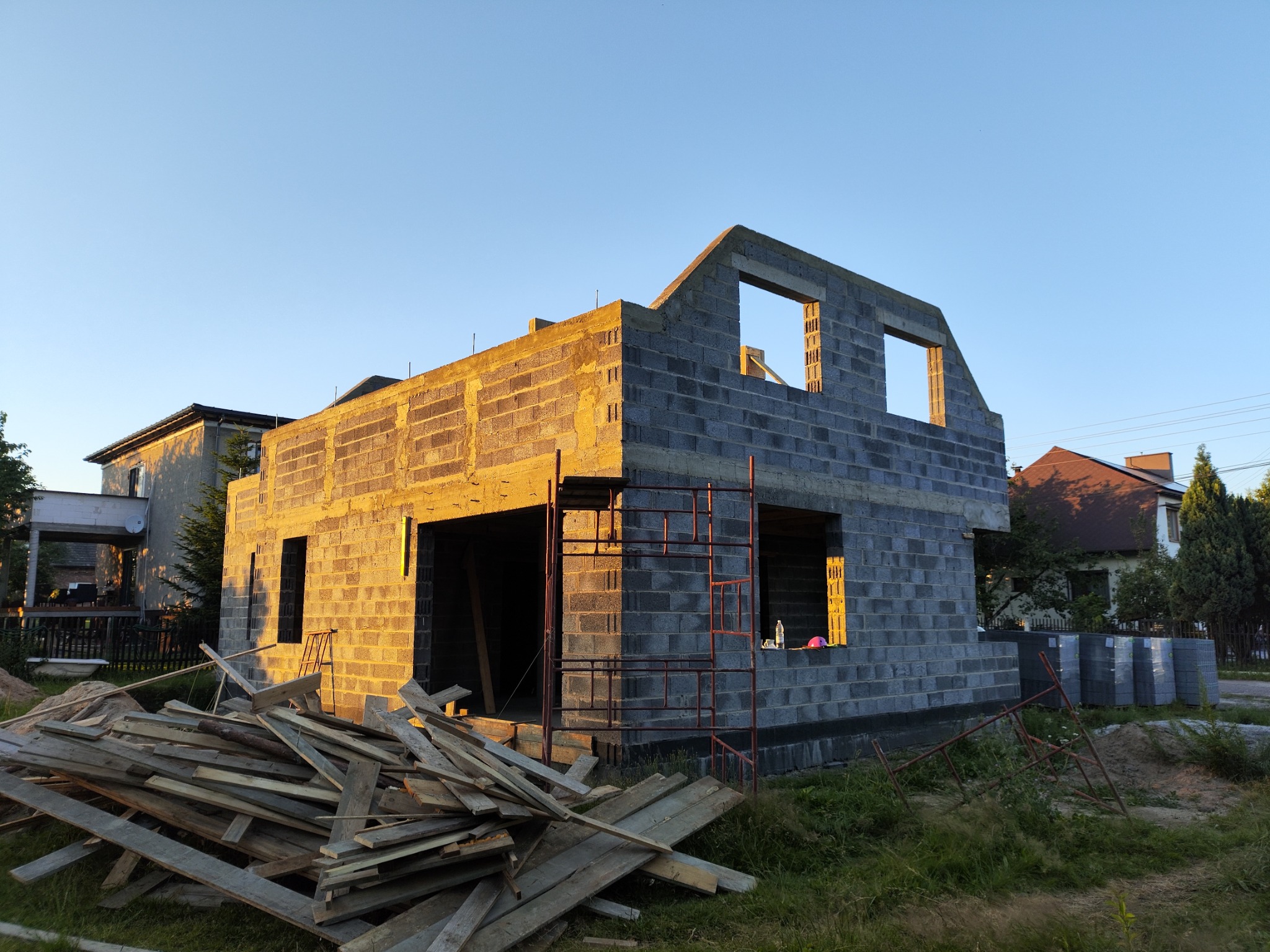 Surowy stan domu z bloczków, wczesnym rankiem, z widocznymi rusztowaniami i stertą desek na pierwszym planie, przy wschodzącym słońcu.