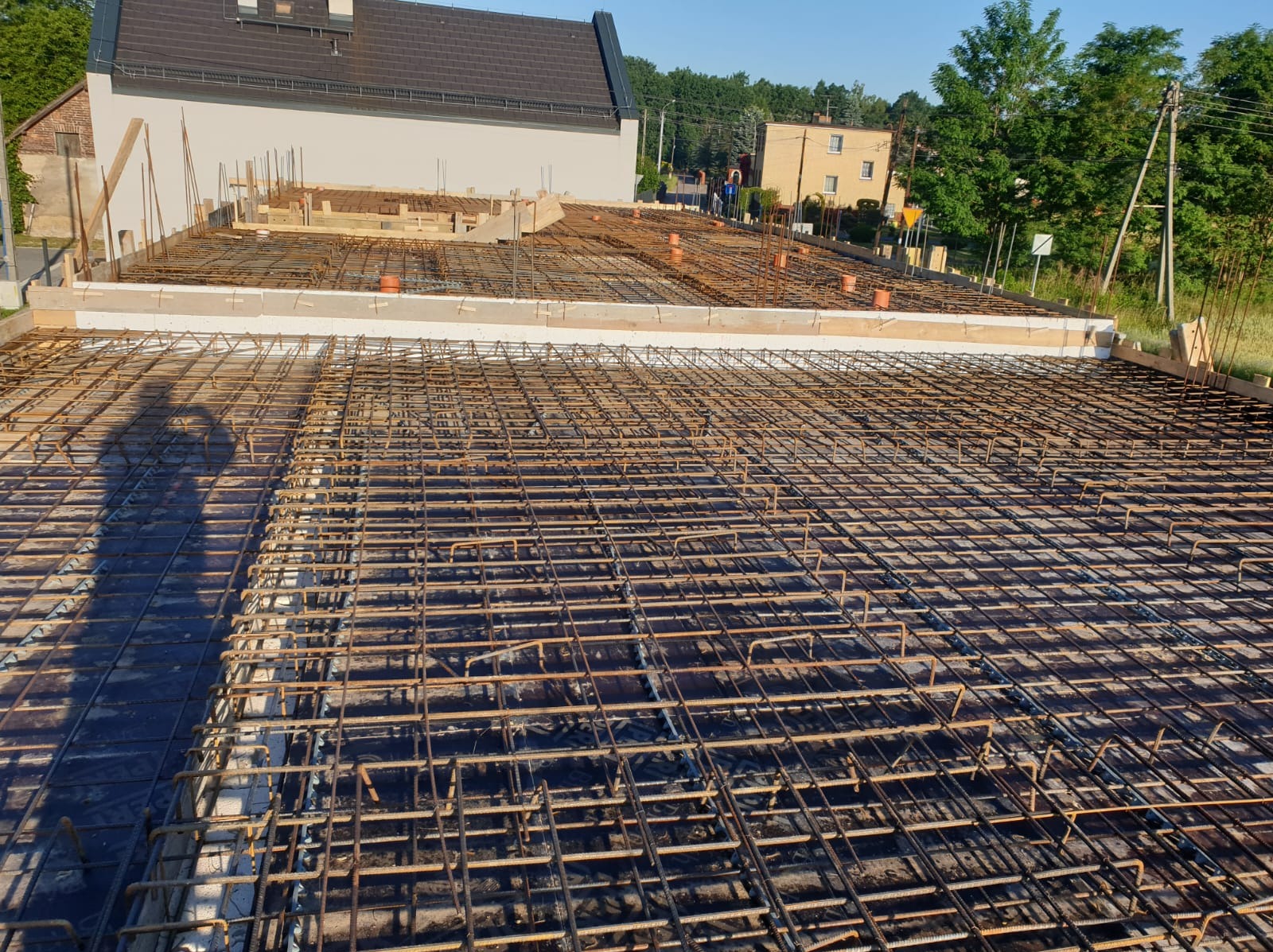 Zbrojenie płyty fundamentowej domu jednorodzinnego, widoczne pręty zbrojeniowe ułożone w kratę, szalunek z białego styropianu oraz wystające rury instalacyjne