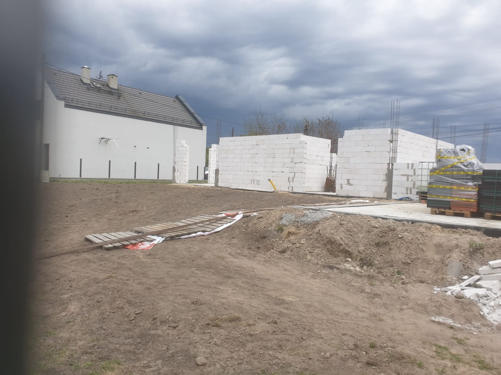 Wznoszone ściany parterowego domu z białych bloczków, widoczne zbrojenia i paleta z materiałami budowlanymi na placu budowy w pochmurny dzień.