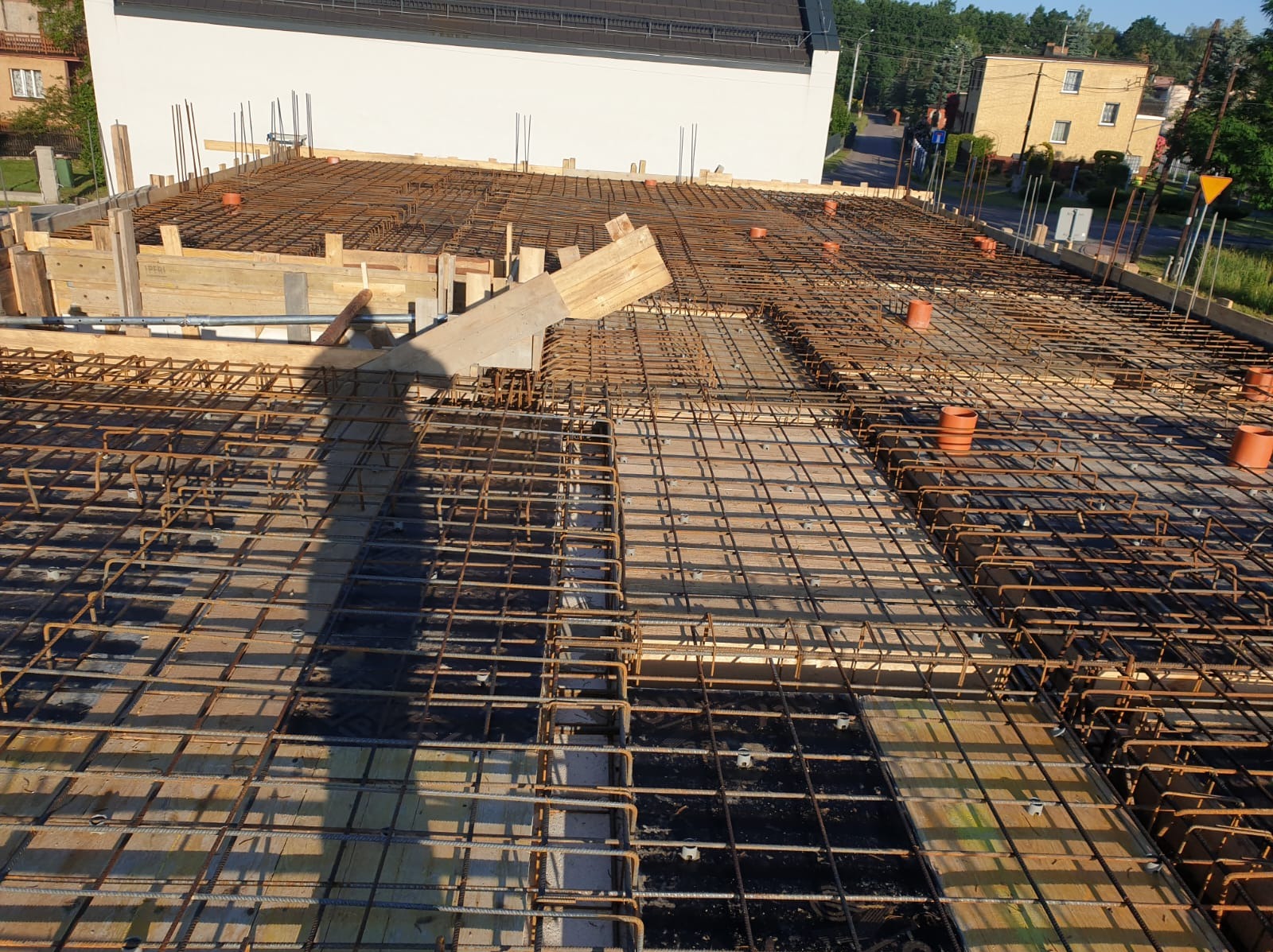 Zbrojenie płyty fundamentowej z deskowaniem i wystającymi prętami, widok z góry na plac budowy z widocznymi budynkami w tle.
