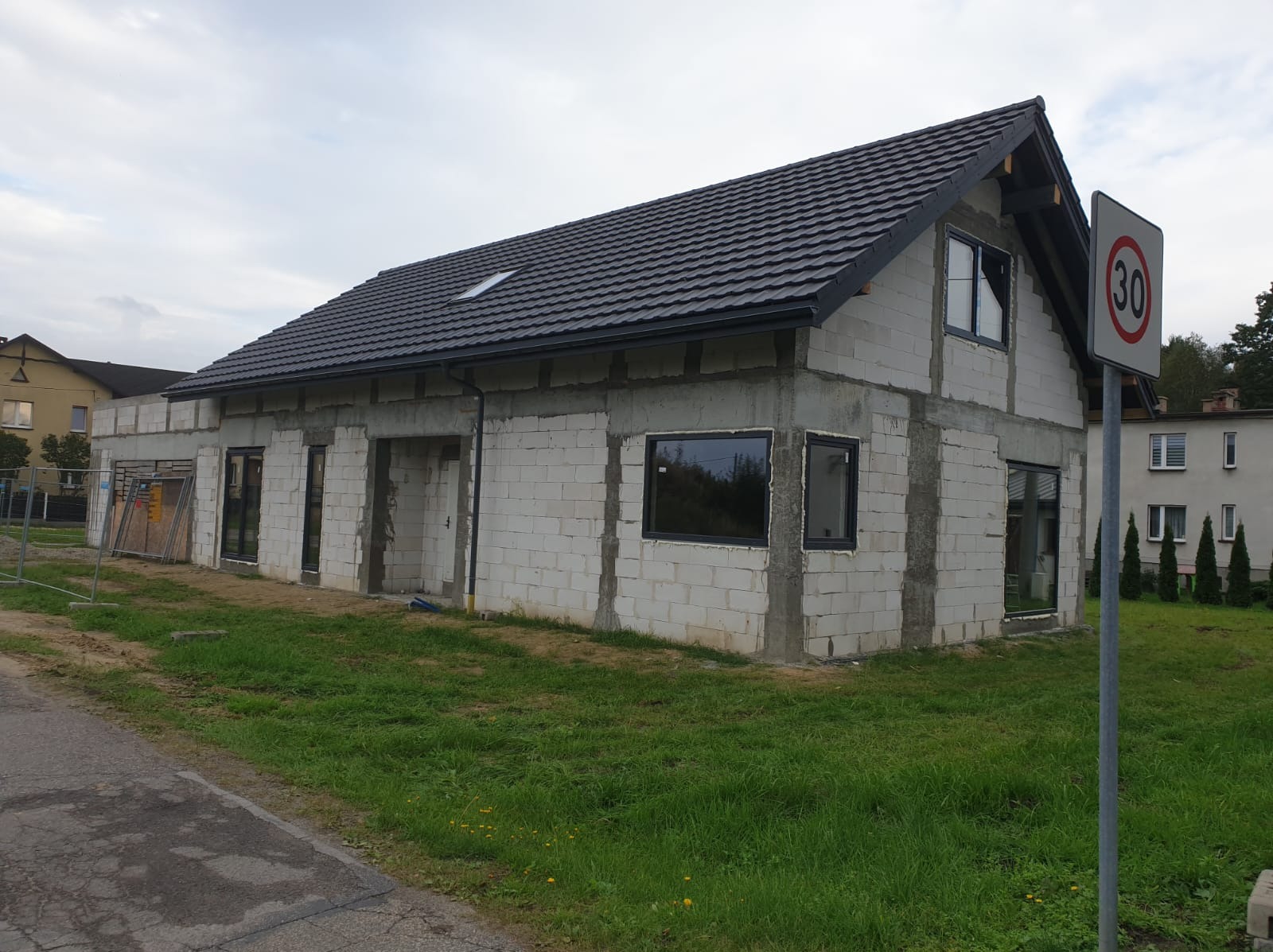 Surowy stan domu z białych bloczków, czarne okna, dach z ciemnej dachówki, znak drogowy ograniczenia prędkości 30 km/h z prawej strony.
