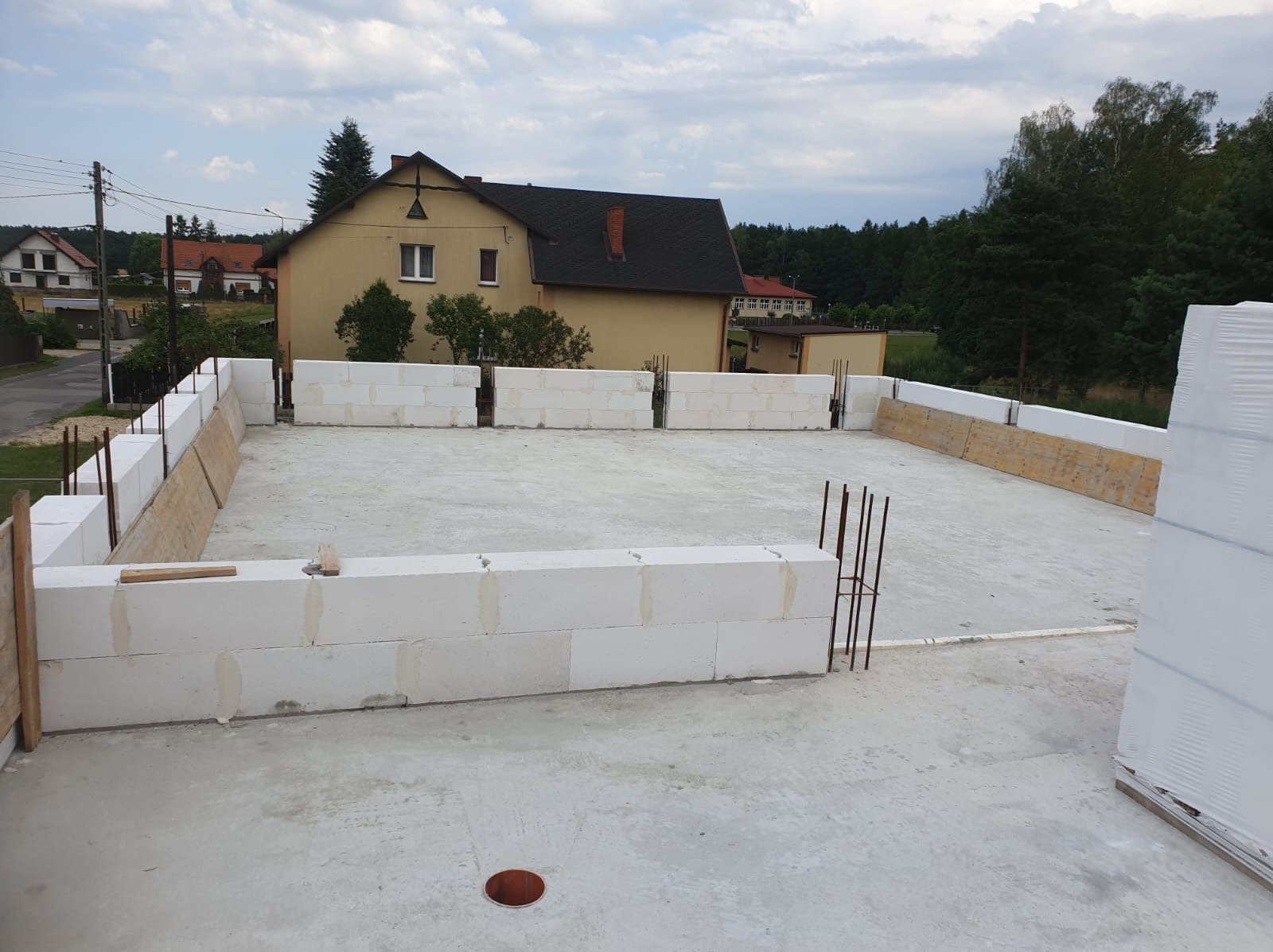 Wznoszenie ścian parteru z betonu komórkowego na płycie fundamentowej, widoczne pręty zbrojeniowe oraz szalunki, w tle dom jednorodzinny w zabudowie podmiejskiej.
