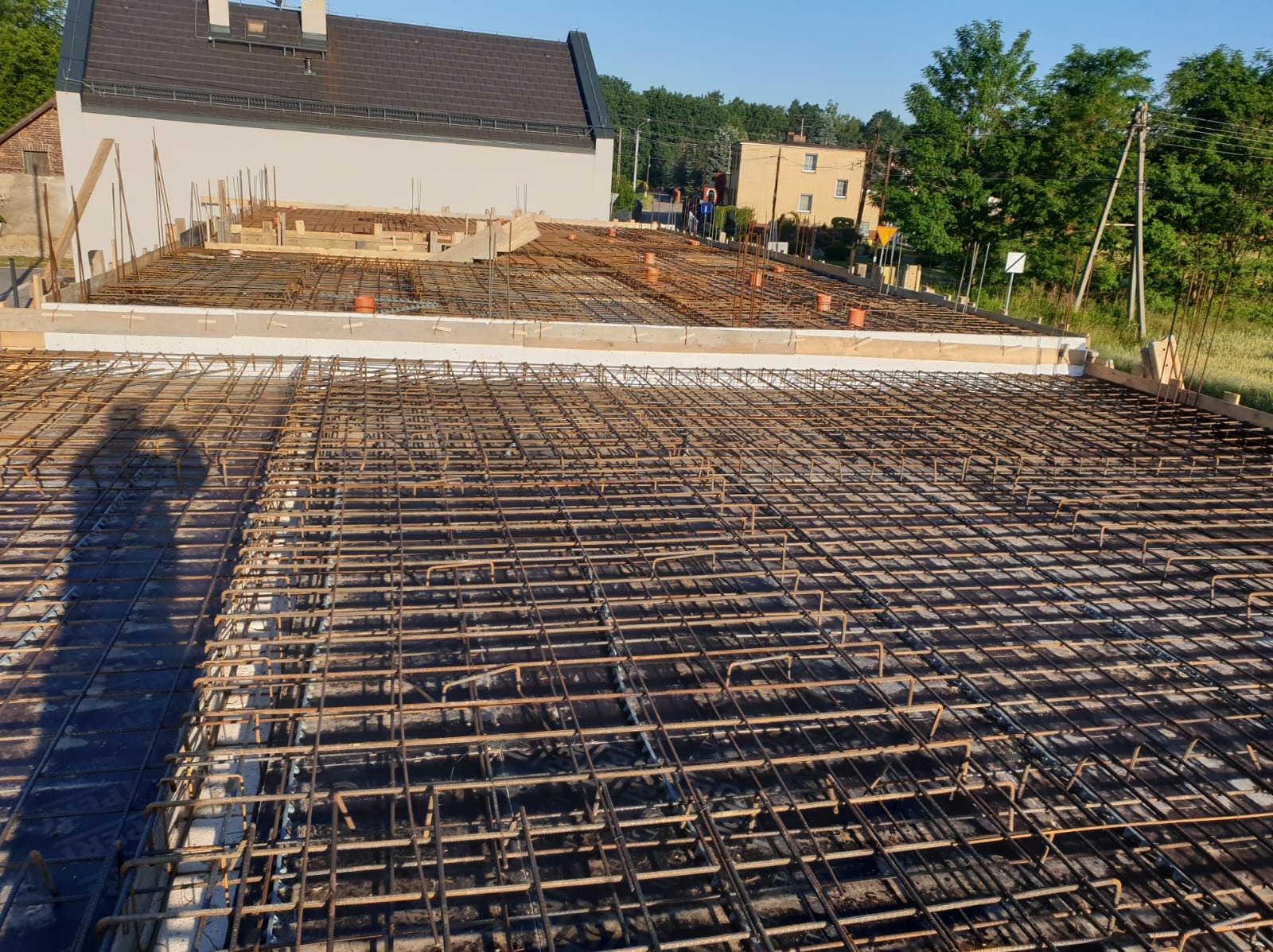 Zbrojenie płyty fundamentowej z widocznymi elementami szalunku i wystającymi prętami, w tle zabudowa jednorodzinna i zieleń.