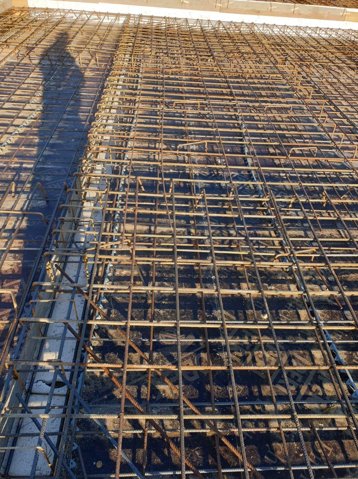 Siatka zbrojeniowa ułożona na deskowaniu, przygotowana do zalania betonem, z widocznym cieniem osoby fotografującej.