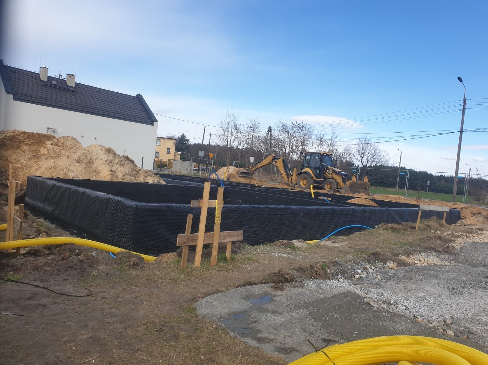 Żółta koparka zasypuje piaskiem wykop pod budynek, zabezpieczony czarną folią. Widoczne wystające rury i drewniane stemple. W tle dom i drzewa.
