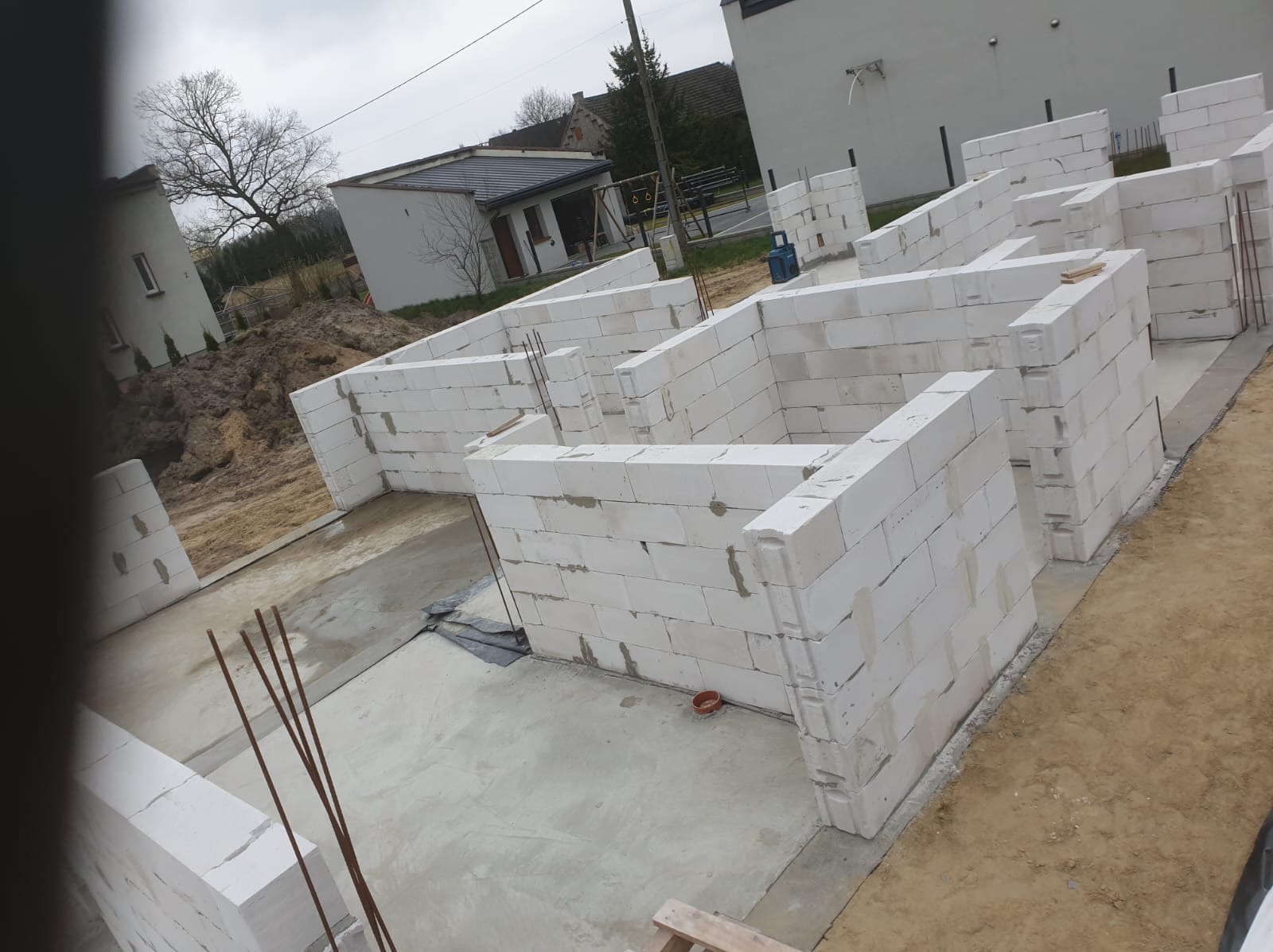 Wznoszenie ścian fundamentowych z białych bloczków na wylanym fundamencie, widoczne wystające pręty zbrojeniowe i otaczający teren budowy.