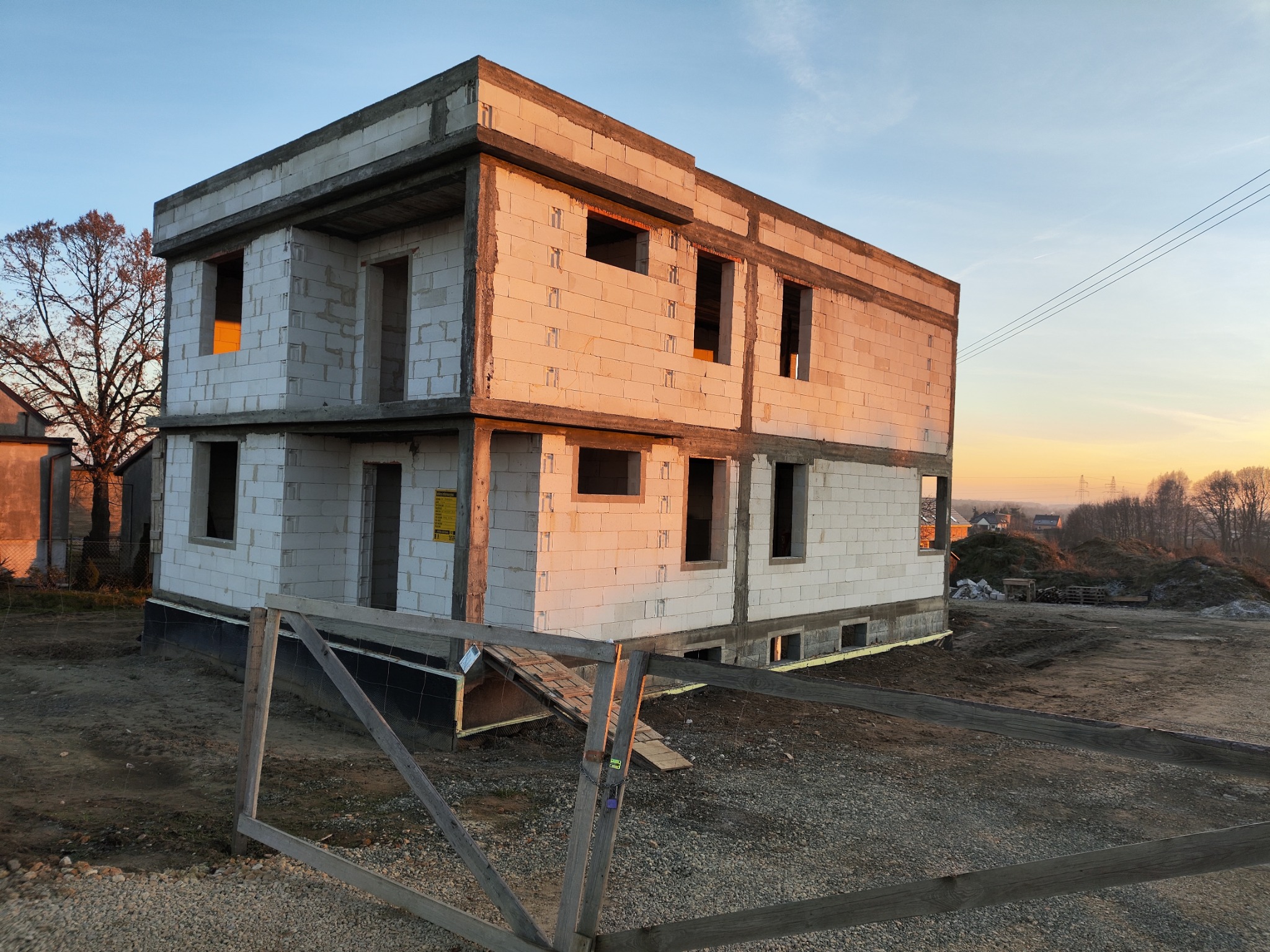 Surowy dwukondygnacyjny budynek z białych bloczków w Bieruniu, widoczna izolacja fundamentów i tymczasowe ogrodzenie na tle zachodzącego słońca.