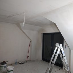 CeramicBud - Poddasze w Namysłowie po szpachlowaniu, ściany i sufit na biało, aluminiowa drabina Krause, widoczne puszki z farbą i akcesoria malarskie, okno balkonowe z czarną ramą.