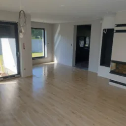 Salon w trakcie wykańczania z ułożoną podłogą z paneli imitujących drewno, widoczne niedokończone ściany, okna z roletami zewnętrznymi i nowoczesny kominek z drewnem wewnątrz.