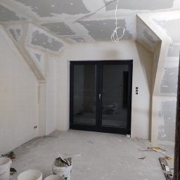 CeramicBud - Pokój w trakcie remontu: ściany i sufit po szpachlowaniu, widoczne płyty kartonowo-gipsowe, przygotowane do malowania, na podłodze wiadra z zaprawą i narzędzia.