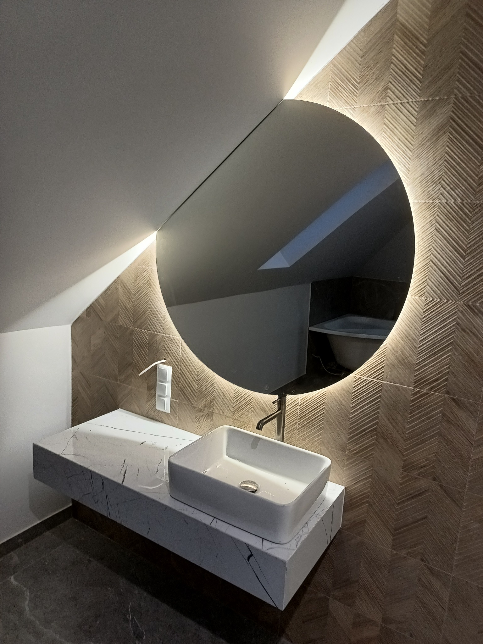 Nowoczesna umywalka nablatowa o kwadratowym kształcie, zamontowana na marmurowym blacie w łazience na poddaszu. W tle okrągłe lustro z podświetleniem LED i ściana wyłożona płytkami w jodełkę.