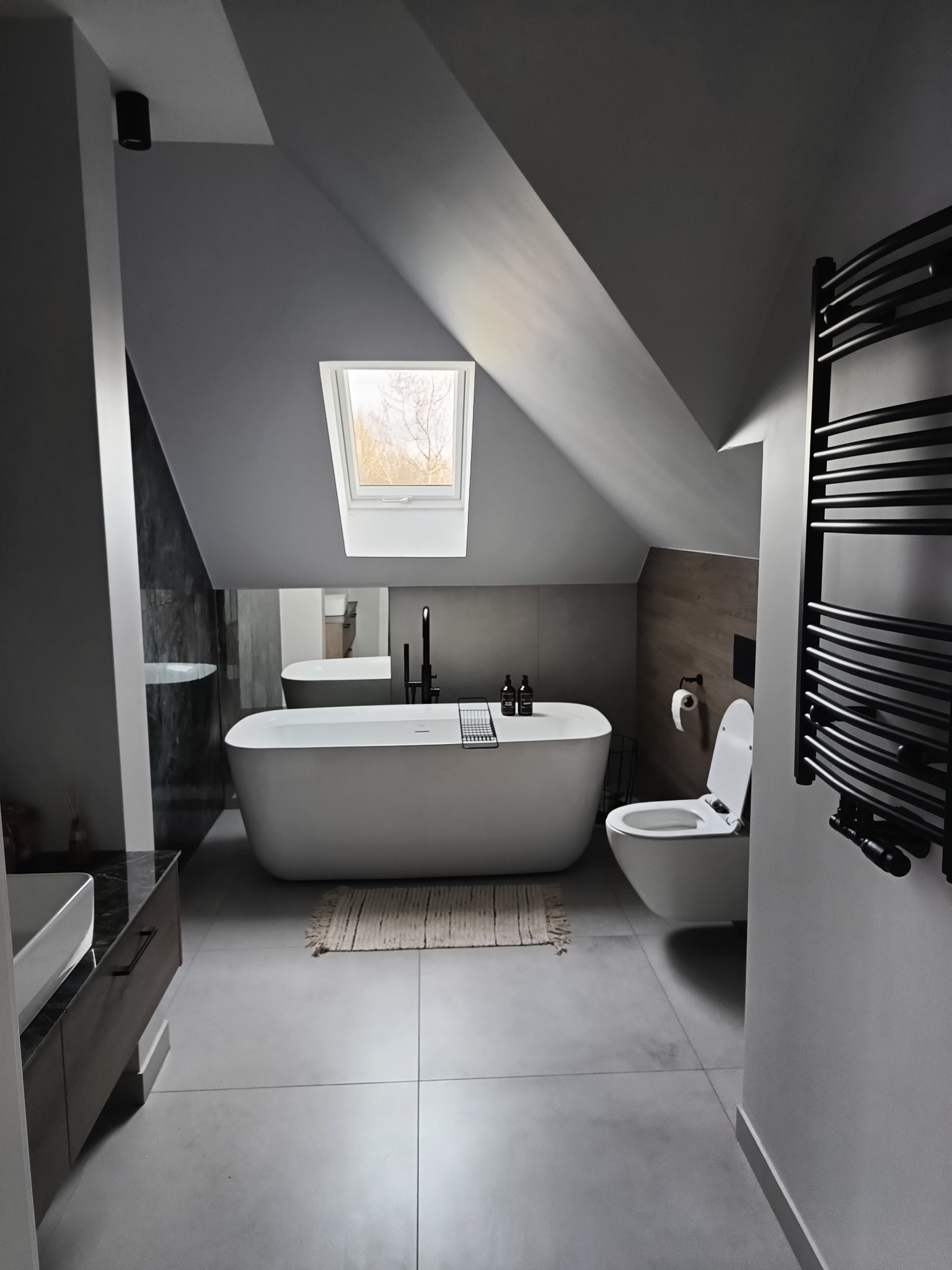 Nowoczesna łazienka na poddaszu ze skośnym sufitem, wyposażona w wolnostojącą wannę, wiszącą toaletę, grzejnik drabinkowy w kolorze czarnym i okno dachowe z widokiem na drzewa.