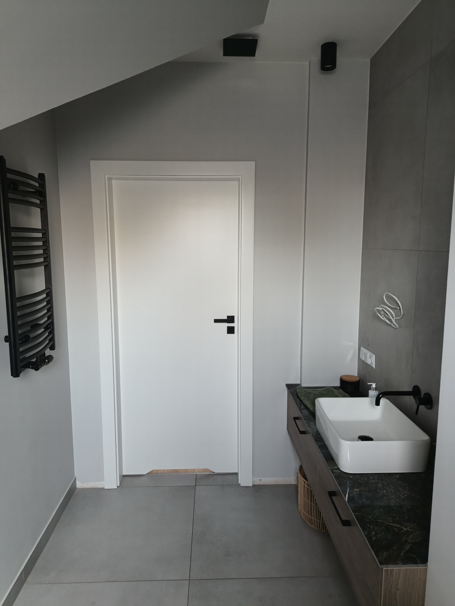 Nowoczesna łazienka na poddaszu z szarą płytką podłogową, białą umywalką nablatową na szafce z ciemnym blatem, czarną armaturą i grzejnikiem drabinkowym, oraz białymi drzwiami z czarną klamką.
