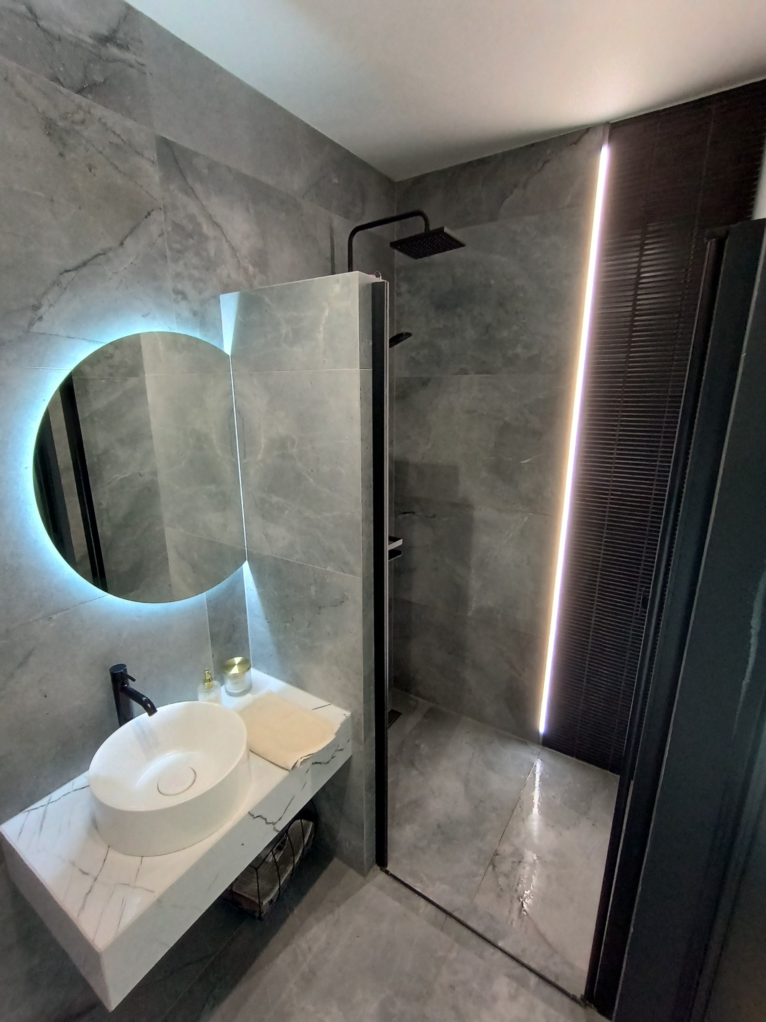 Nowoczesna łazienka z szarymi marmurowymi płytkami, okrągłym lustrem z podświetleniem LED, białą umywalką nablatową i czarną armaturą, kabina prysznicowa typu walk-in z czarnymi drzwiami.
