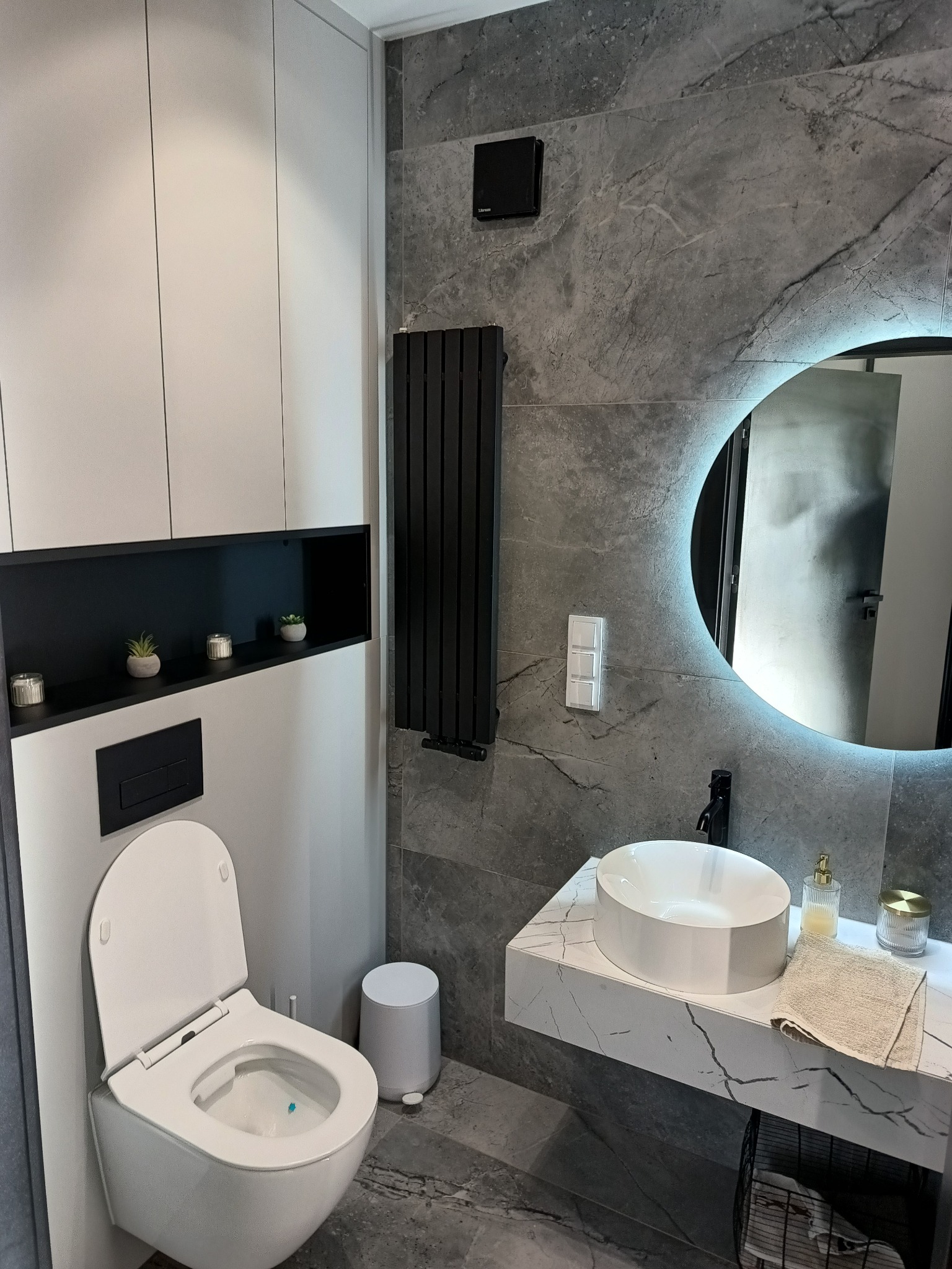 Nowoczesna łazienka z wiszącą toaletą, umywalką nablatową na marmurowym blacie, podświetlanym okrągłym lustrem i grzejnikiem drabinkowym na ścianie z szarych płytek imitujących marmur.