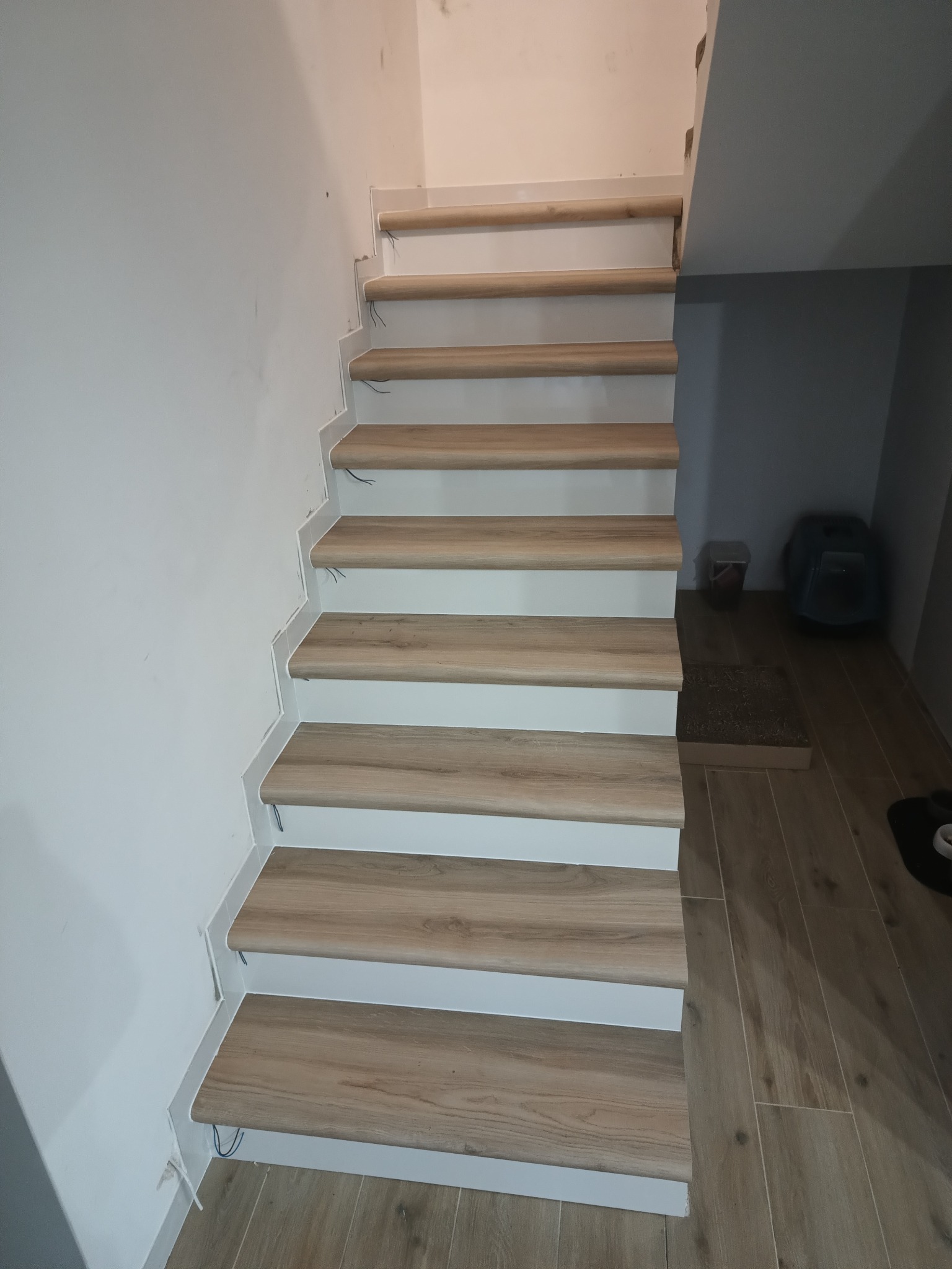 Wykończone schody wewnętrzne z drewnopodobnymi panelami, białe podstopnice, widoczne kable wystające ze ściany, podłoga wyłożona panelami pod kolor schodów.