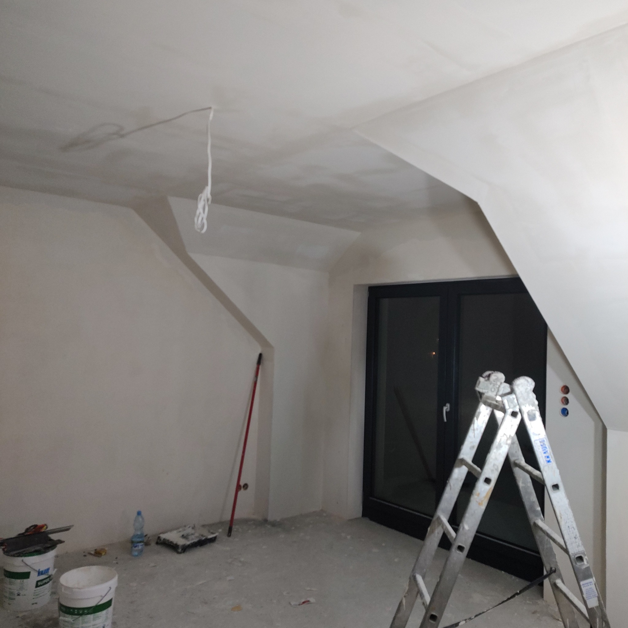 Poddasze w Namysłowie po szpachlowaniu, ściany i sufit na biało, aluminiowa drabina Krause, widoczne puszki z farbą i akcesoria malarskie, okno balkonowe z czarną ramą.