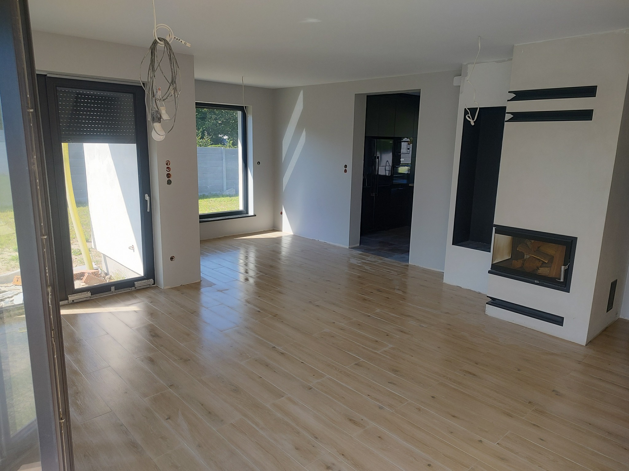 Salon w trakcie wykańczania z ułożoną podłogą z paneli imitujących drewno, widoczne niedokończone ściany, okna z roletami zewnętrznymi i nowoczesny kominek z drewnem wewnątrz.