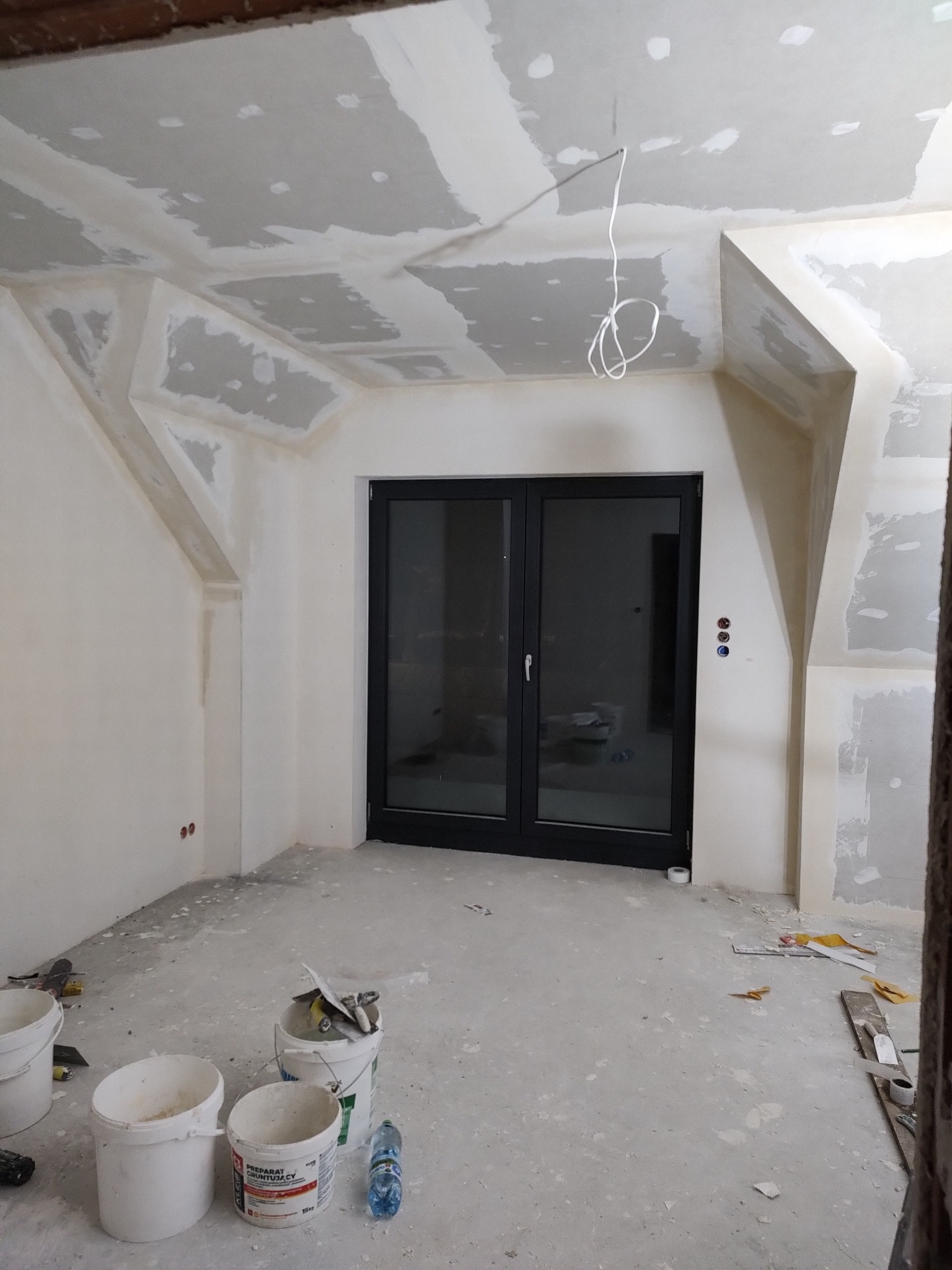 Pokój w trakcie remontu: ściany i sufit po szpachlowaniu, widoczne płyty kartonowo-gipsowe, przygotowane do malowania, na podłodze wiadra z zaprawą i narzędzia.