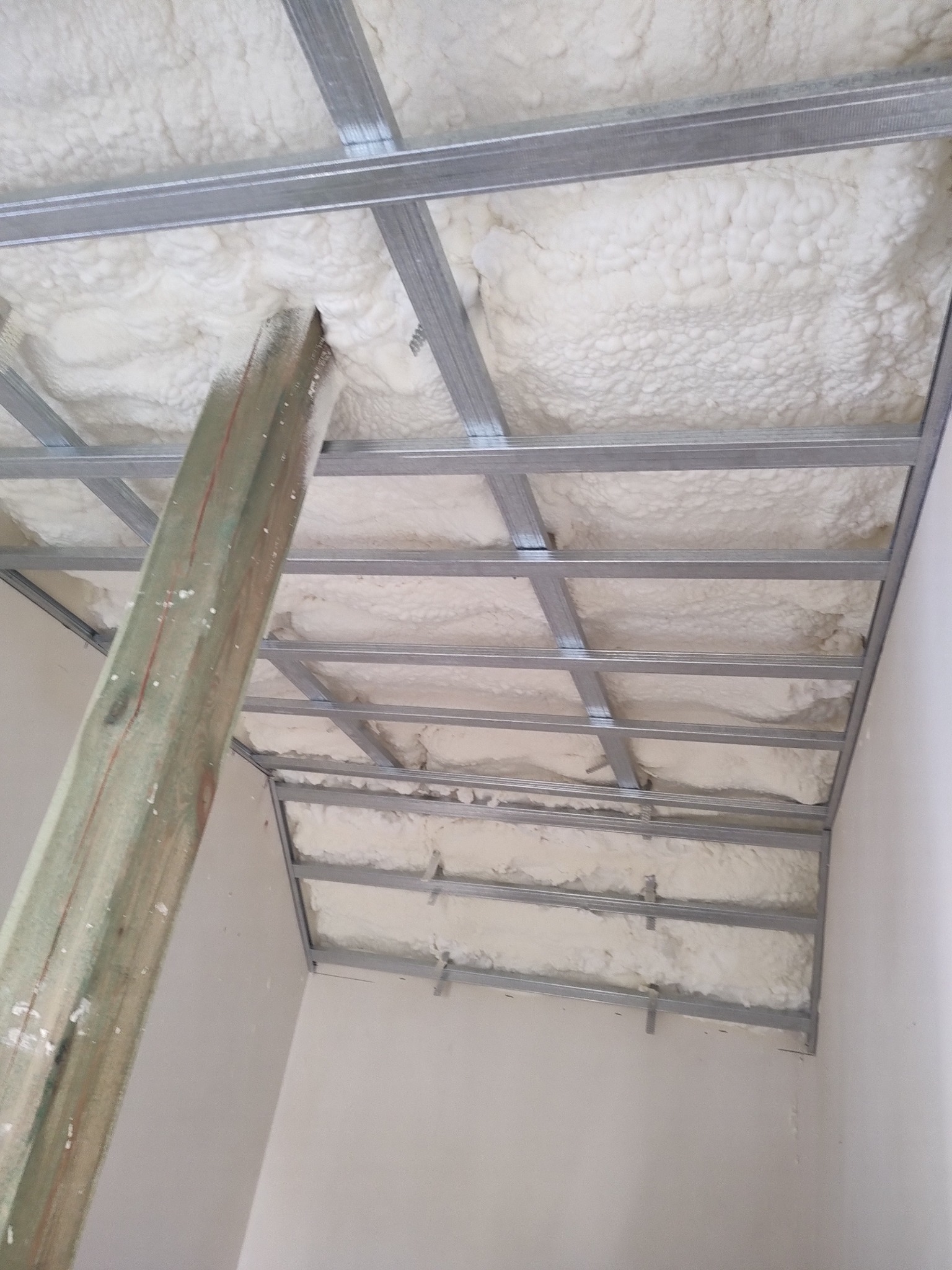 Montaż metalowego stelaża sufitu podwieszanego z wypełnieniem pianką izolacyjną, widoczny drewniany słup podpierający konstrukcję.