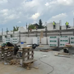 Budowa domu z betonu kom&oacute;rkowego H+H. Murarze w ż&oacute;łtych kamizelkach układają bloczki na ścianie. Na pierwszym planie narzędzia i materiały budowlane.