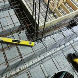 Zbrojenie schodów z użyciem bloczków betonowych, stalowych prętów, drewnianego szalunku i poziomicy. Widoczne narzędzia pracy na budowie.