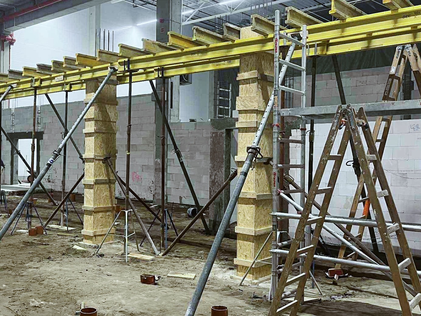Formowanie szalunków kolumnowych z drewnianych płyt na budowie. Widoczne podpory, belki stropowe i ściany z bloczków. W tle drabiny i otwarty stan surowy budynku.