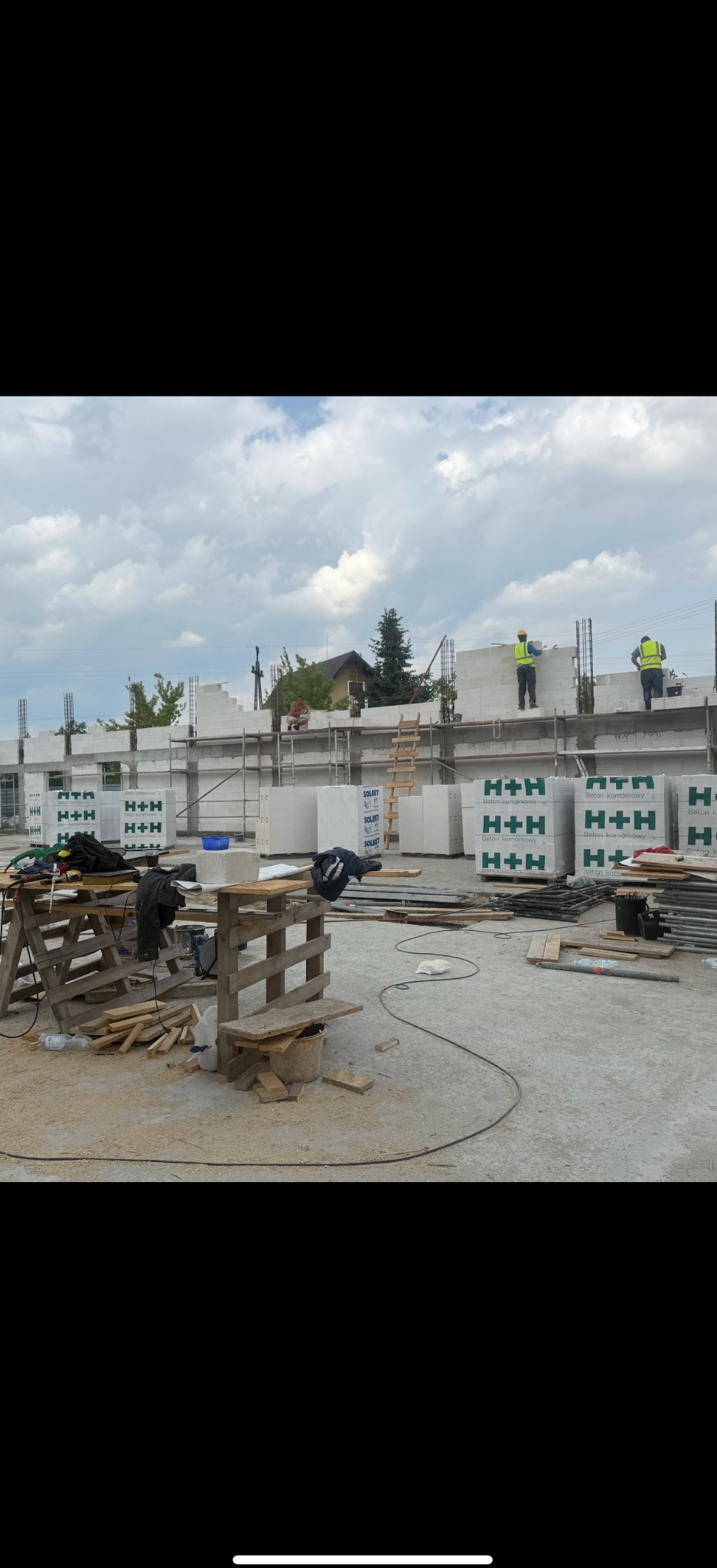 Budowa domu z betonu komórkowego H+H. Murarze w żółtych kamizelkach układają bloczki na ścianie. Na pierwszym planie narzędzia i materiały budowlane.