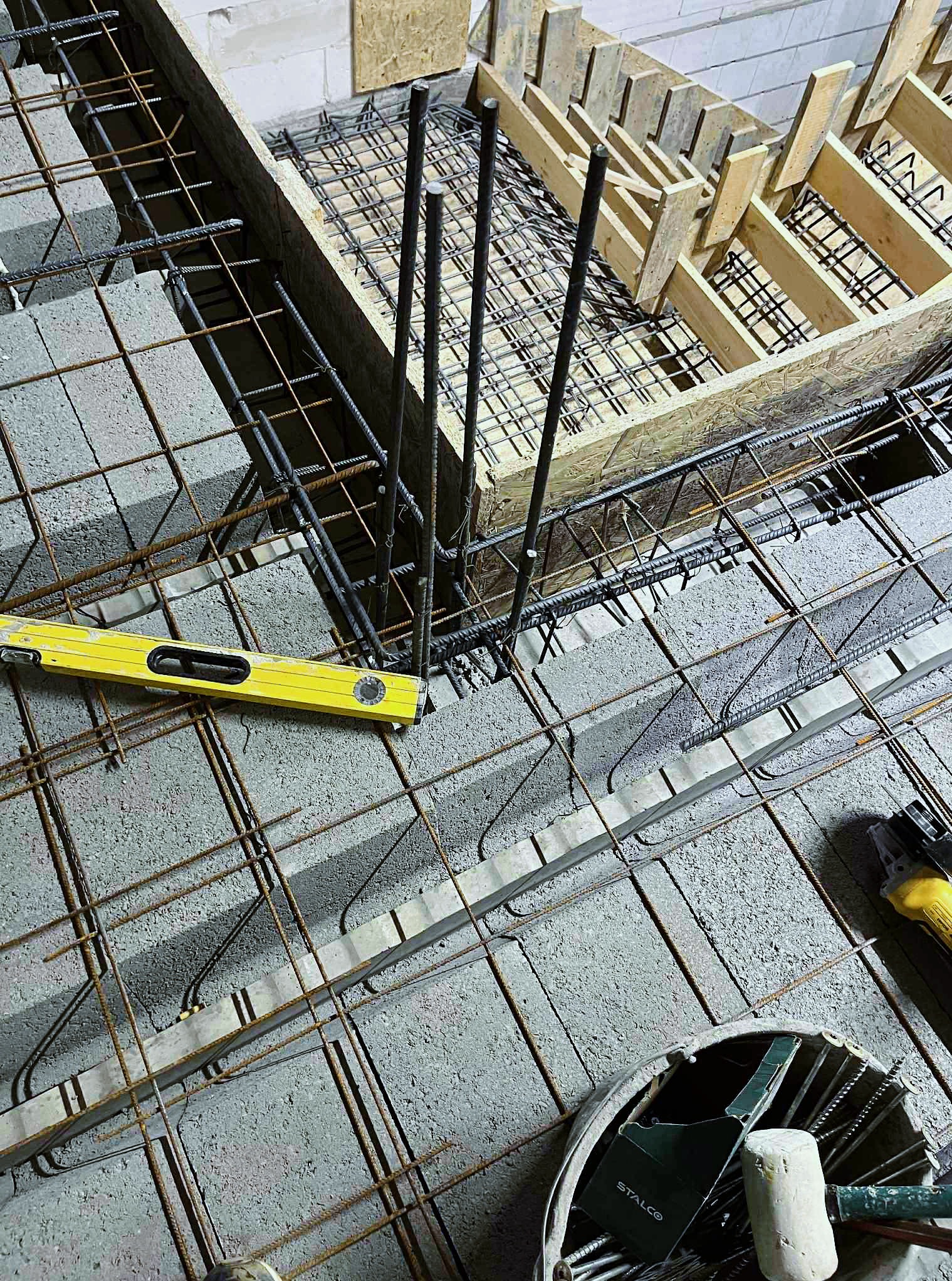 Zbrojenie schodów z użyciem bloczków betonowych, stalowych prętów, drewnianego szalunku i poziomicy. Widoczne narzędzia pracy na budowie.