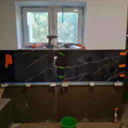 Częściowo wyłożona czarnym marmurem ściana w łazience, widoczne narzędzia, poziomica, szpachla i wiadro, okno w tle, etap prac wykończeniowych.