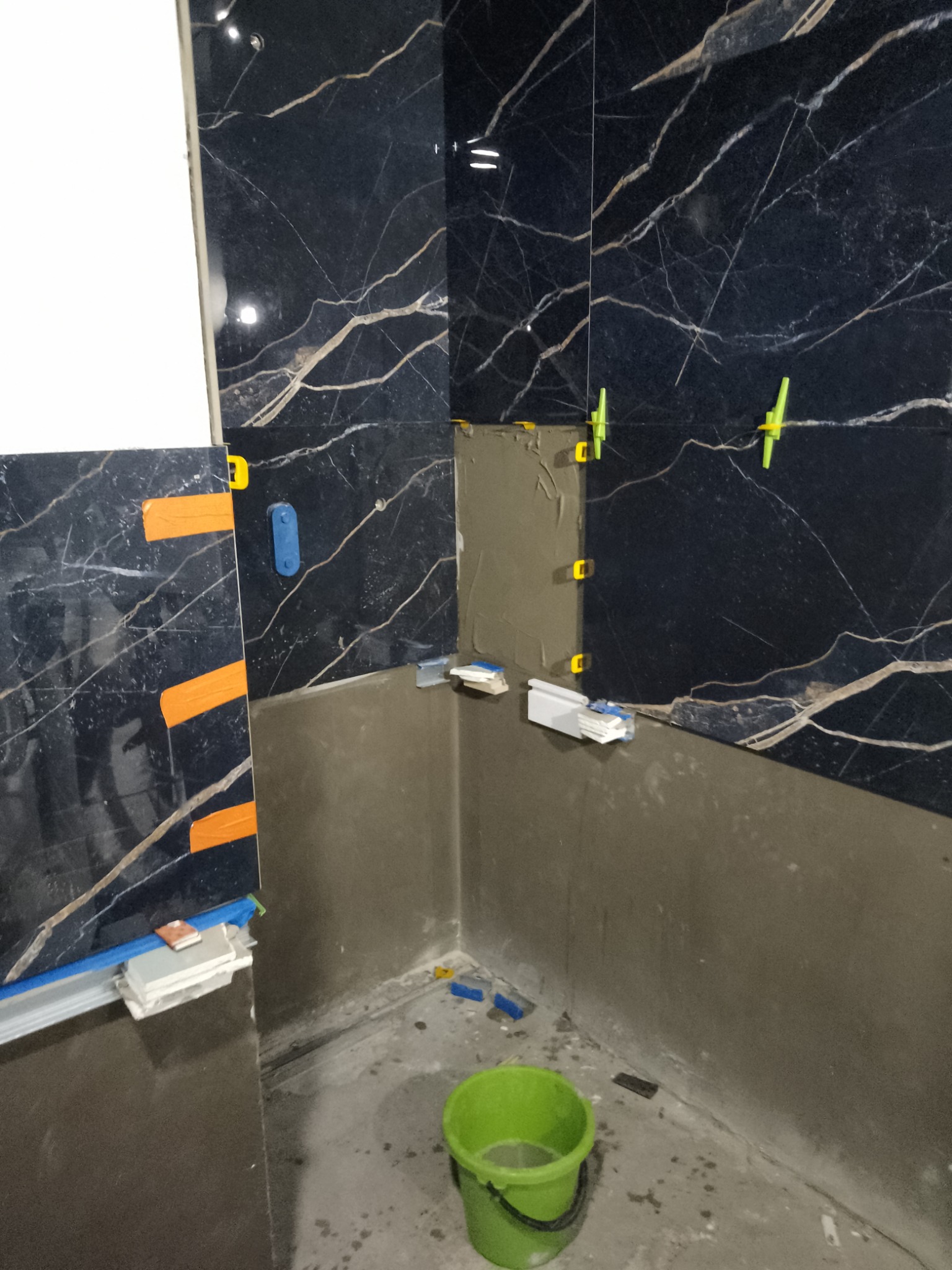 Fragment łazienki w trakcie wykańczania, widoczne czarne płytki z białym żyłkowaniem montowane przy użyciu poziomicy, klipsów i krzyżyków dystansowych, na podłodze zielone wiadro z wodą.