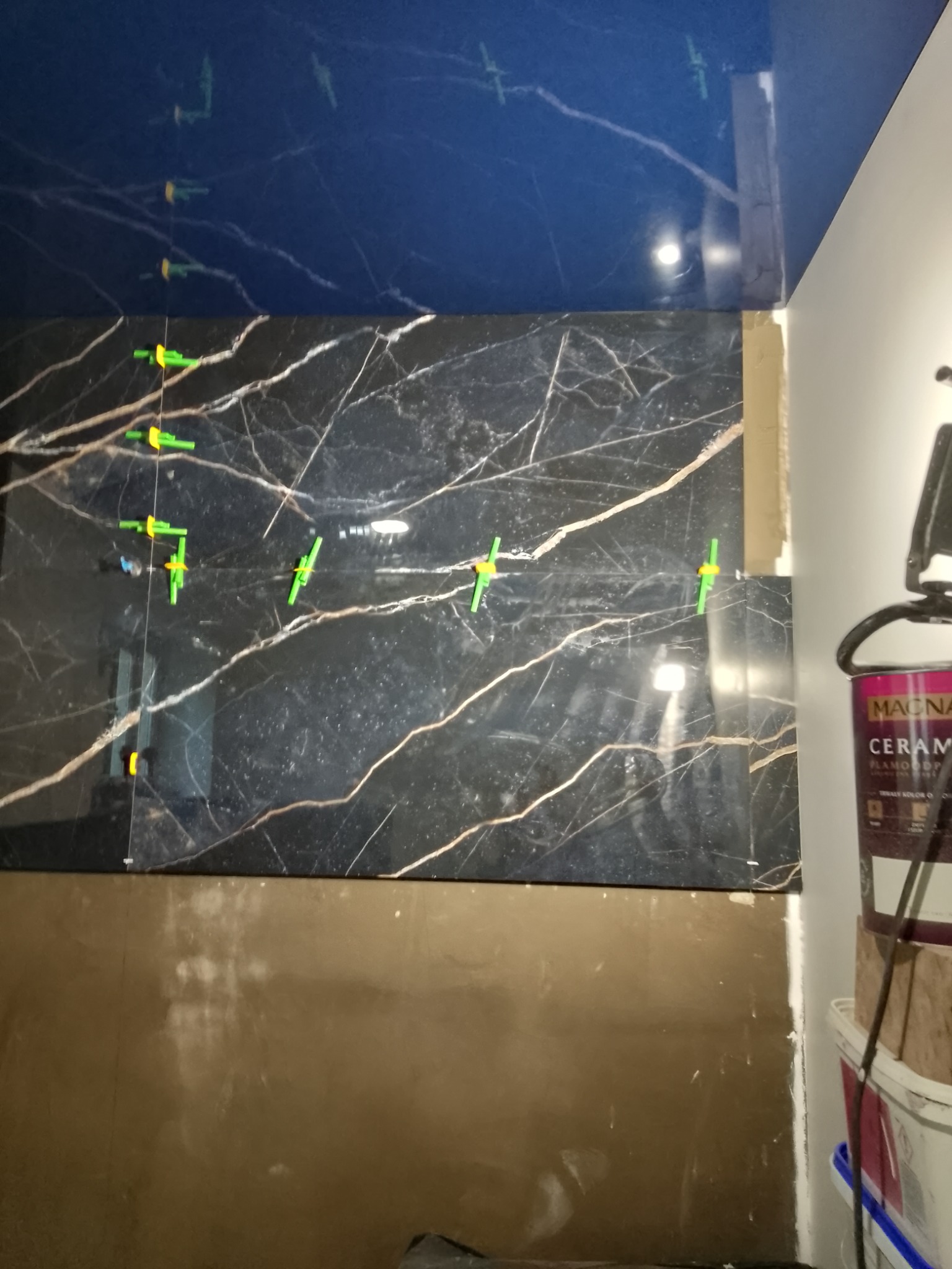 Ściana łazienki w trakcie wykańczania, z położonymi płytkami imitującymi czarny marmur z jasnymi żyłami, użyte zielone krzyżyki dystansowe, widoczny fragment sufitu w kolorze ciemnego granatu.